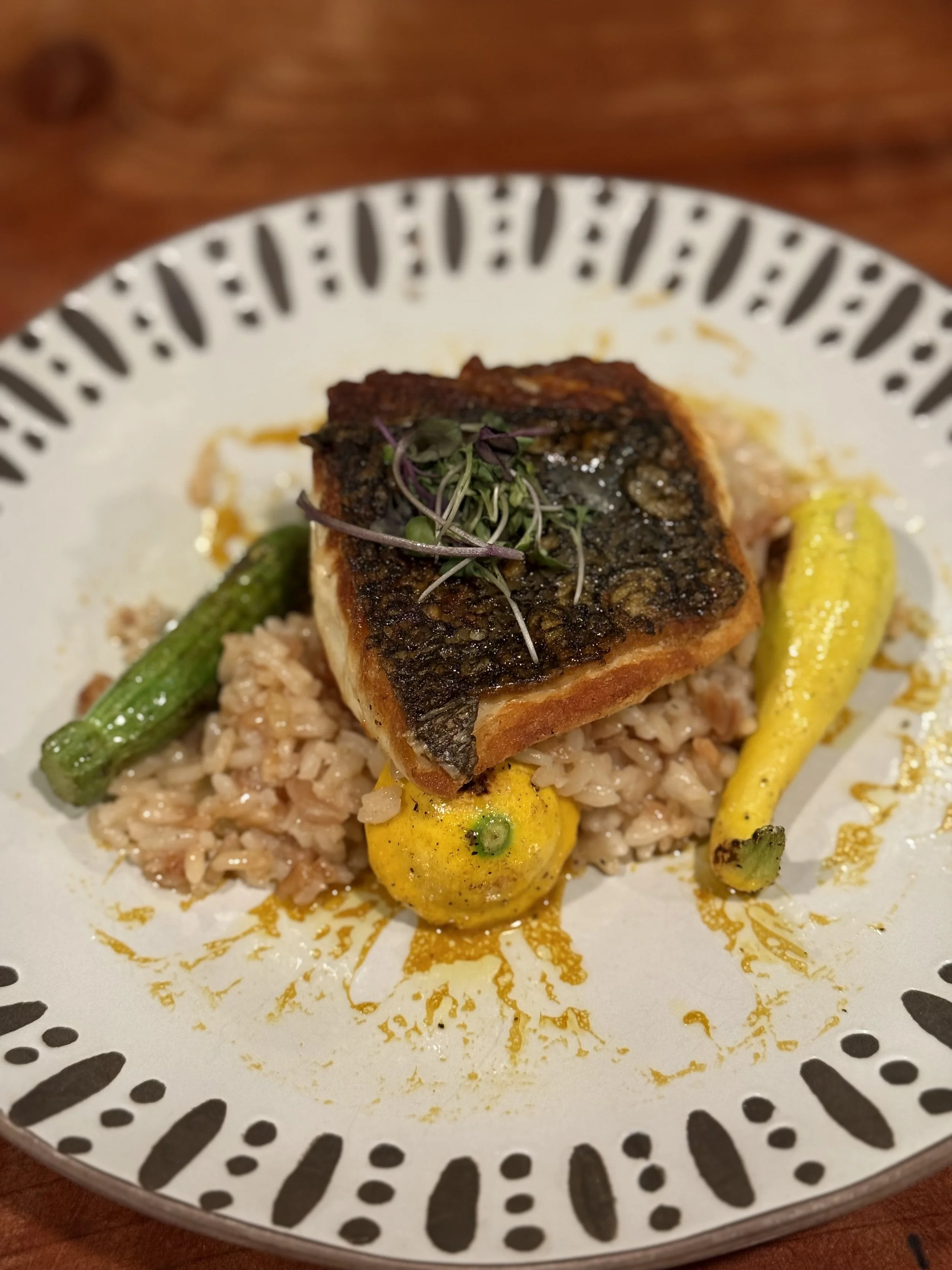 Grilled fish fillet with crispy skin on rice, garnished with microgreens, served with grilled yellow and green chilies on a decorative plate