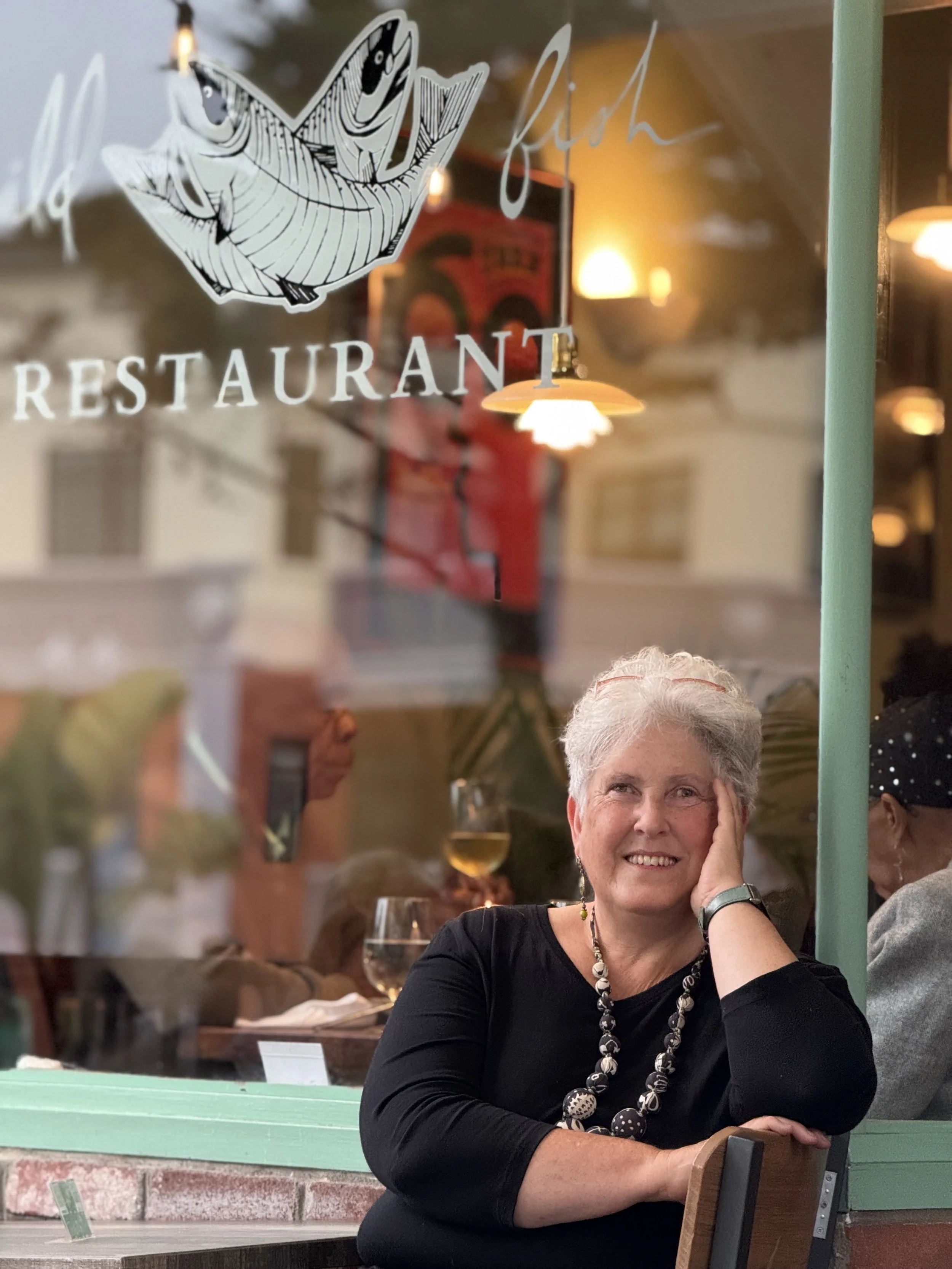 A woman with gray hair sitting outside a restaurant window, smiling, with her hand resting on her face, inside a cozy restaurant with warm lighting and glasses of wine on the table.