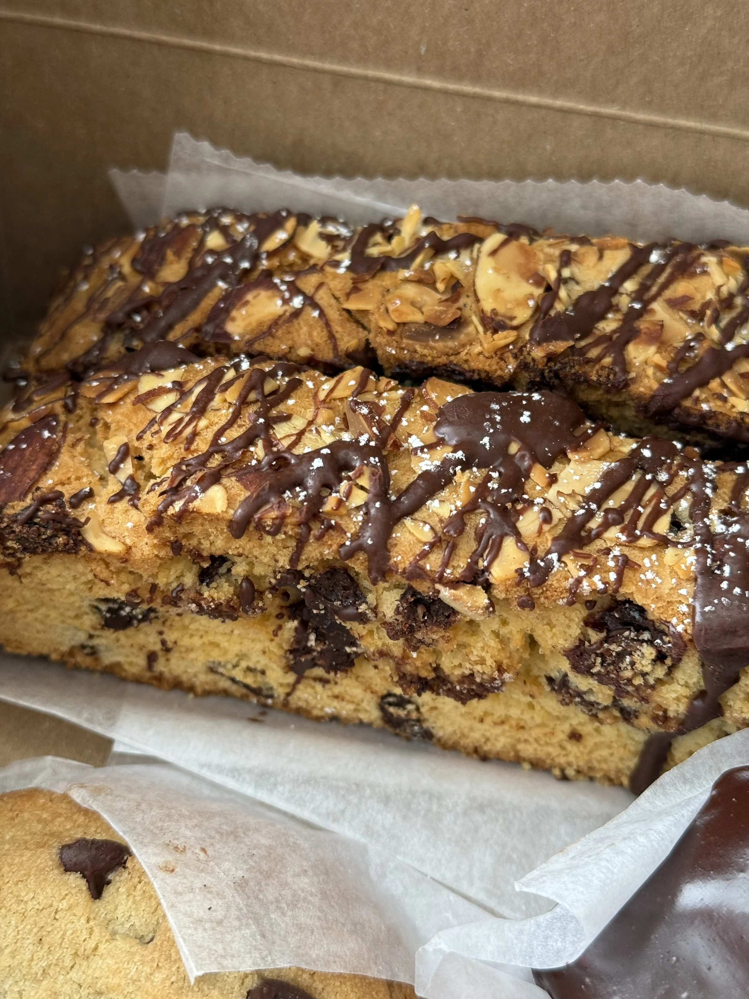 A close-up of a chocolate chip cookie cake topped with almond slices, drizzled with chocolate, and sprinkled with powdered sugar, inside a brown cardboard box.