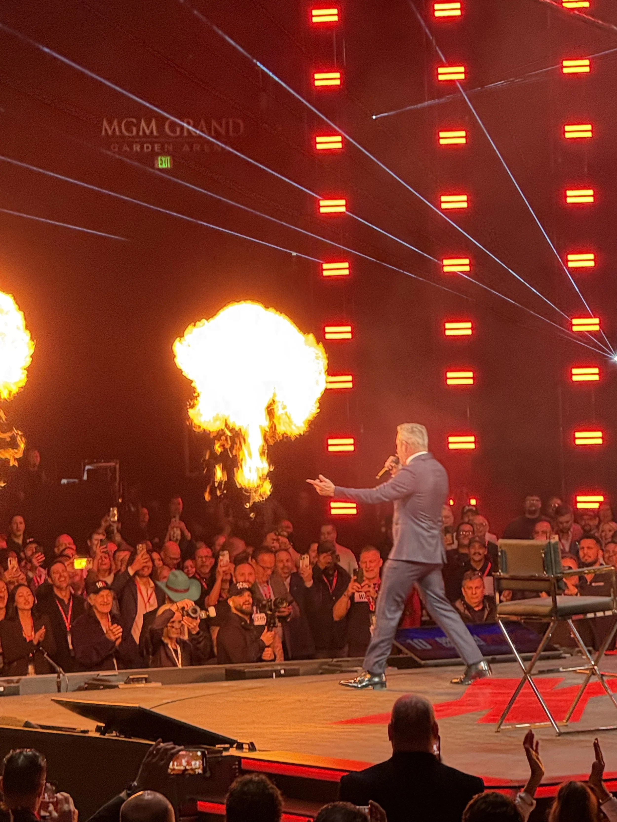 A male speaker in a gray suit performs a fire-breathing act on stage at a conference or event, with flames shooting from his mouth, an audience watching and recording, bright red stage lighting, and laser beams in the background.