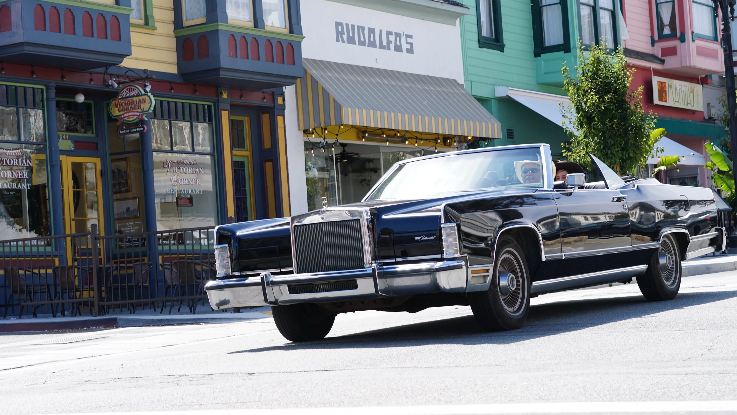 A black convertible classic car driving on a city street with colorful buildings and shops in the background.