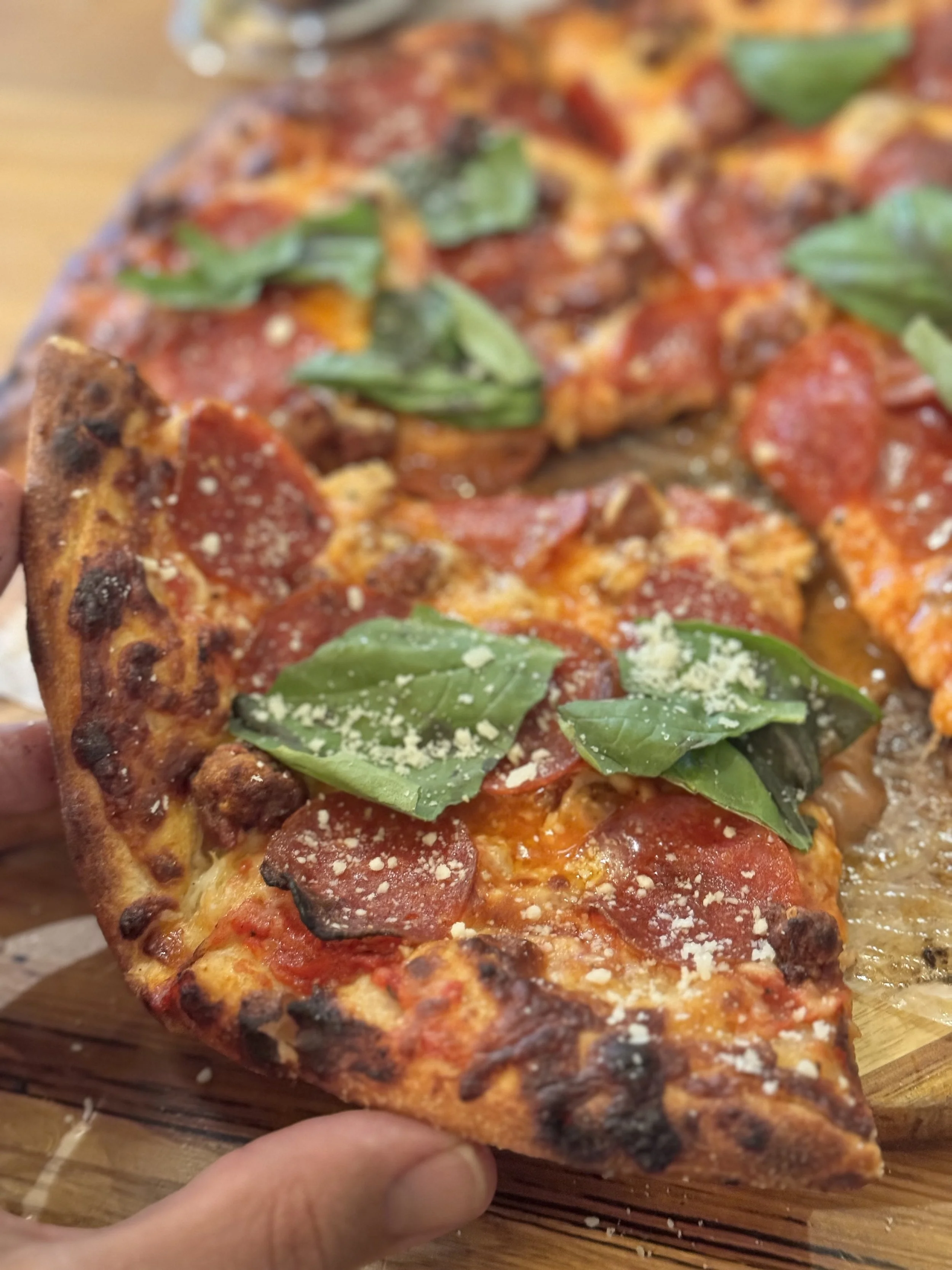 A slice of pepperoni pizza with fresh basil and grated cheese on top, held above a wooden pizza board.