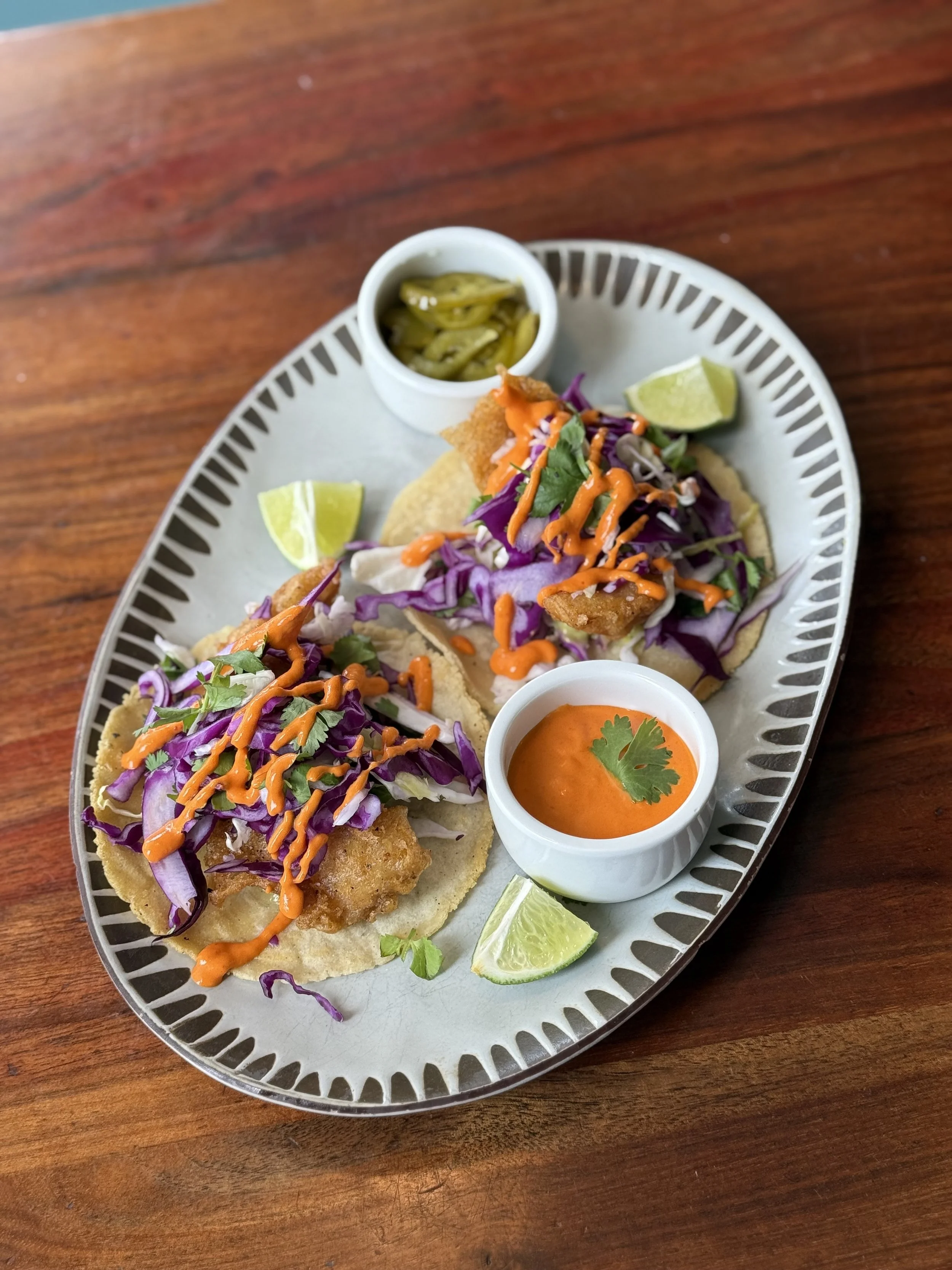 Two fish tacos topped with purple cabbage slaw, drizzled with orange sauce, served with lime wedges, a small bowl of green pickled peppers, and a small bowl of red sauce, all on a gray platter on a wooden table.
