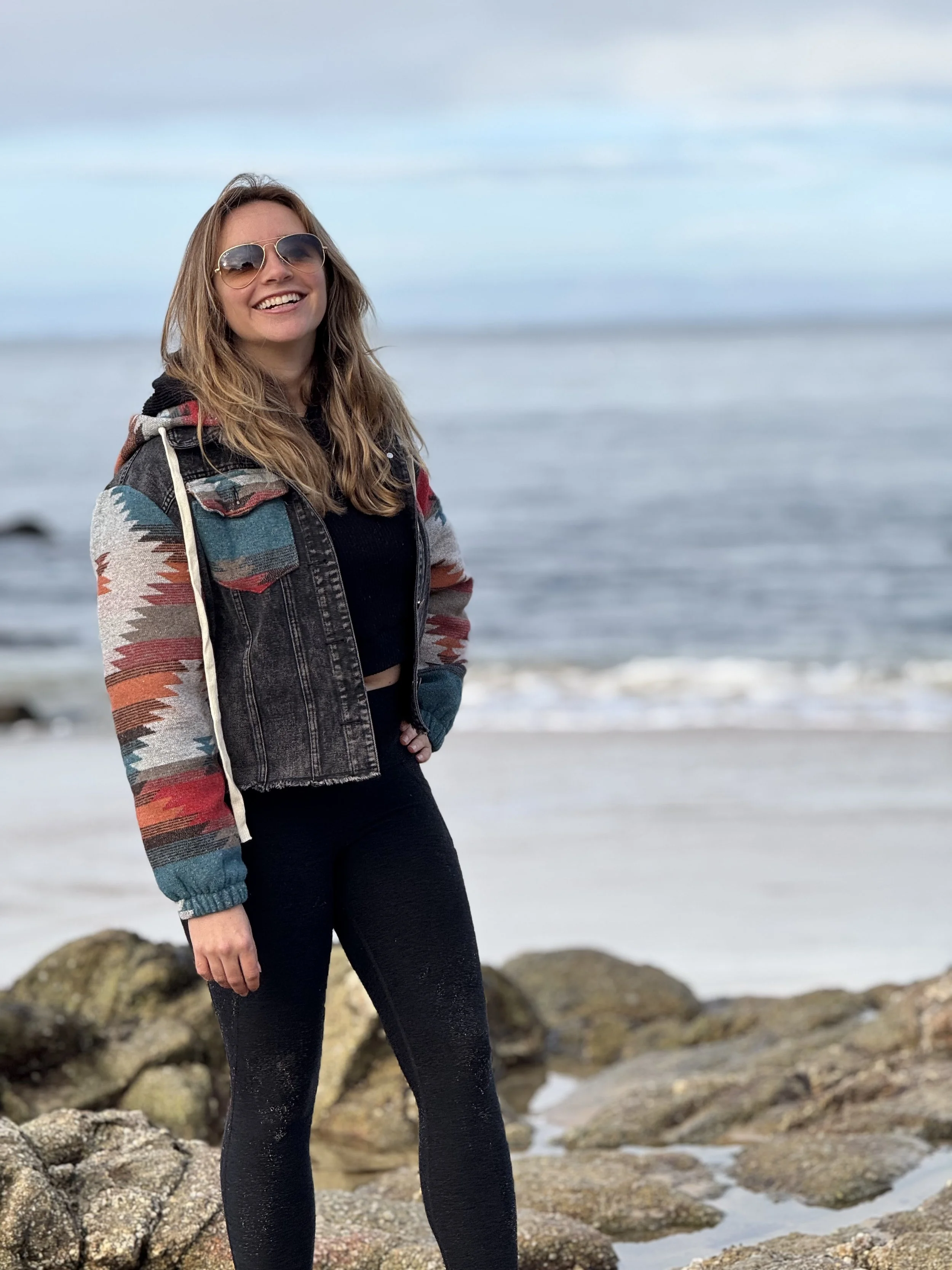 A young woman with long, wavy hair, wearing sunglasses, a colorful patterned jacket, a black top, and black leggings, standing on rocks near the ocean with a partly cloudy sky in the background.