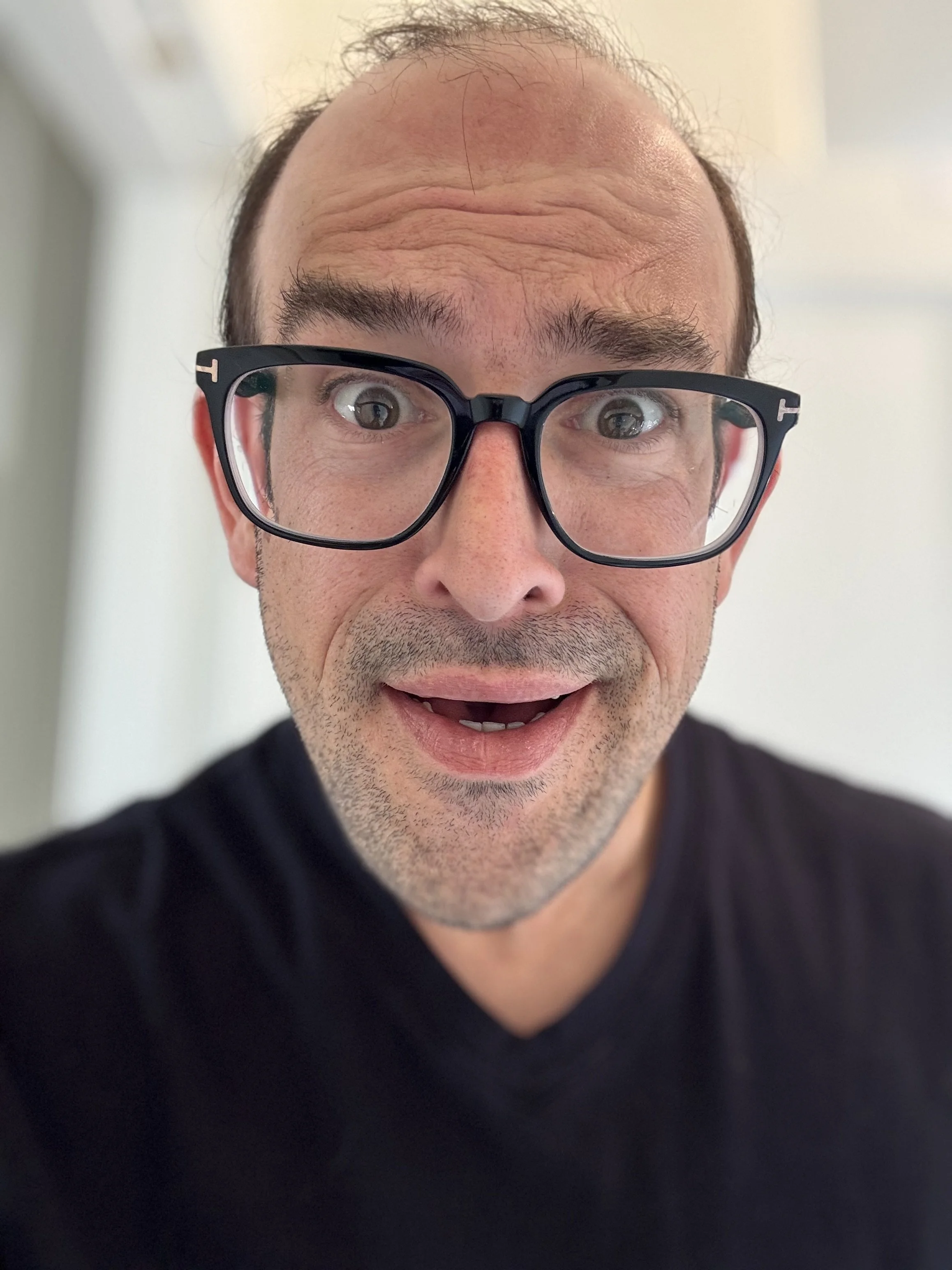 Close-up portrait of a man with glasses and a surprised expression, mouth open, short hair, wearing a black shirt.