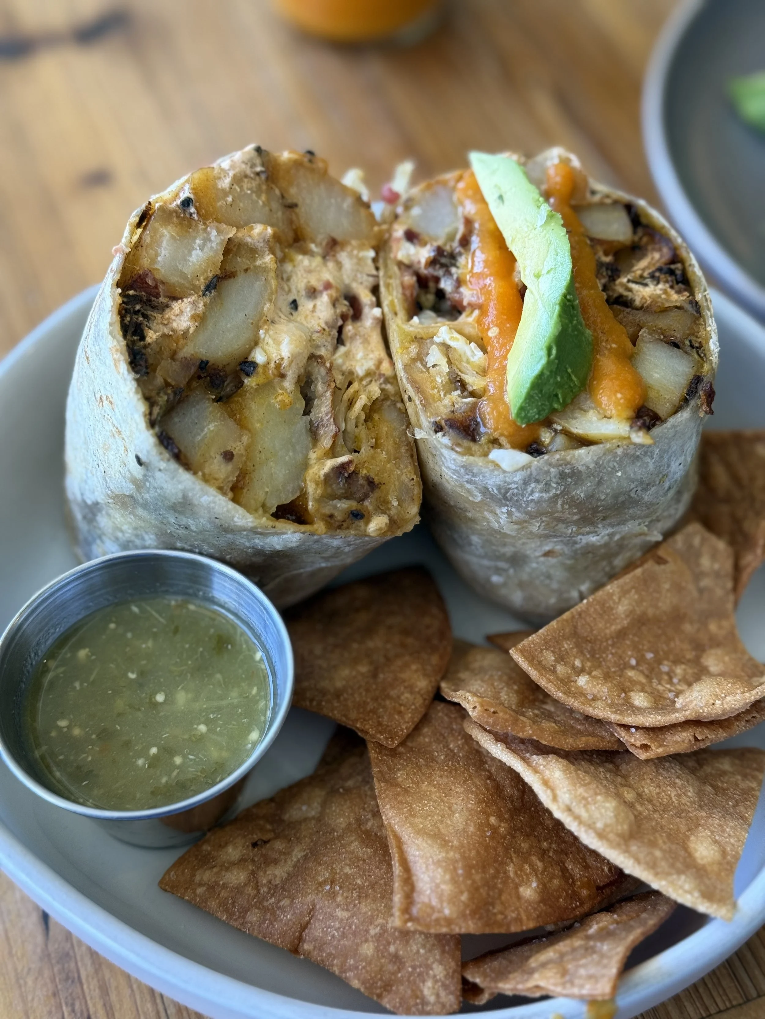 Two breakfast burritos with potato filling, topped with avocado and hot sauce, served with salsa and crispy chips.