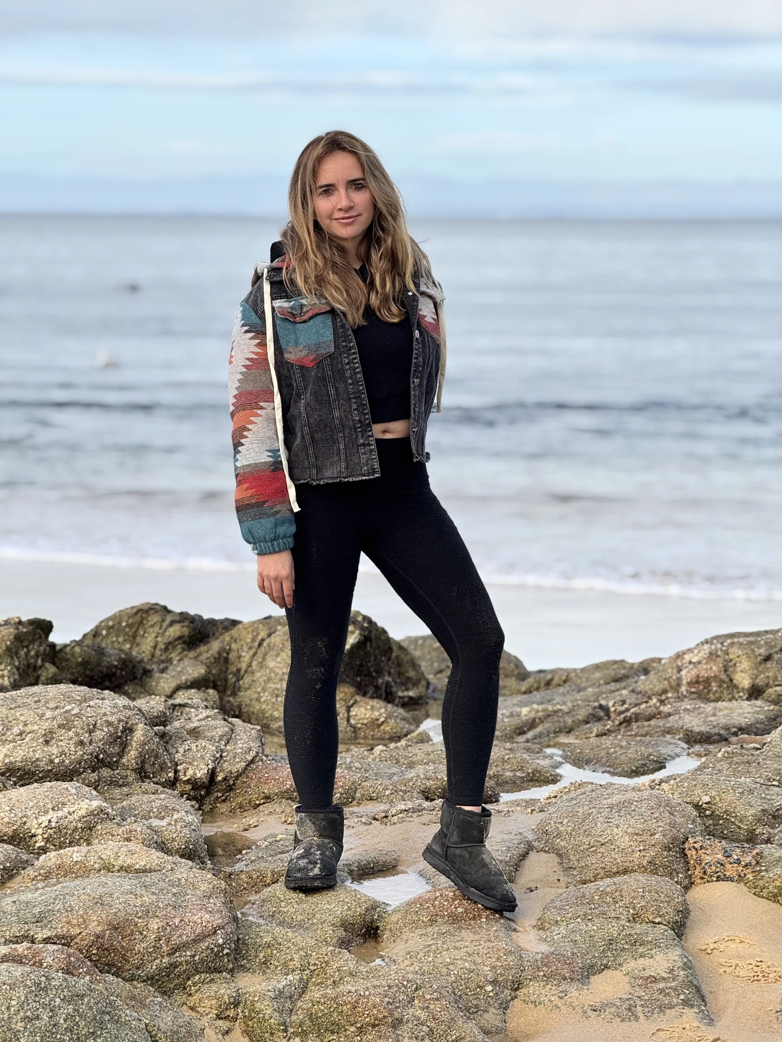 Young woman standing on rocks at the beach with the ocean and sky in the background.