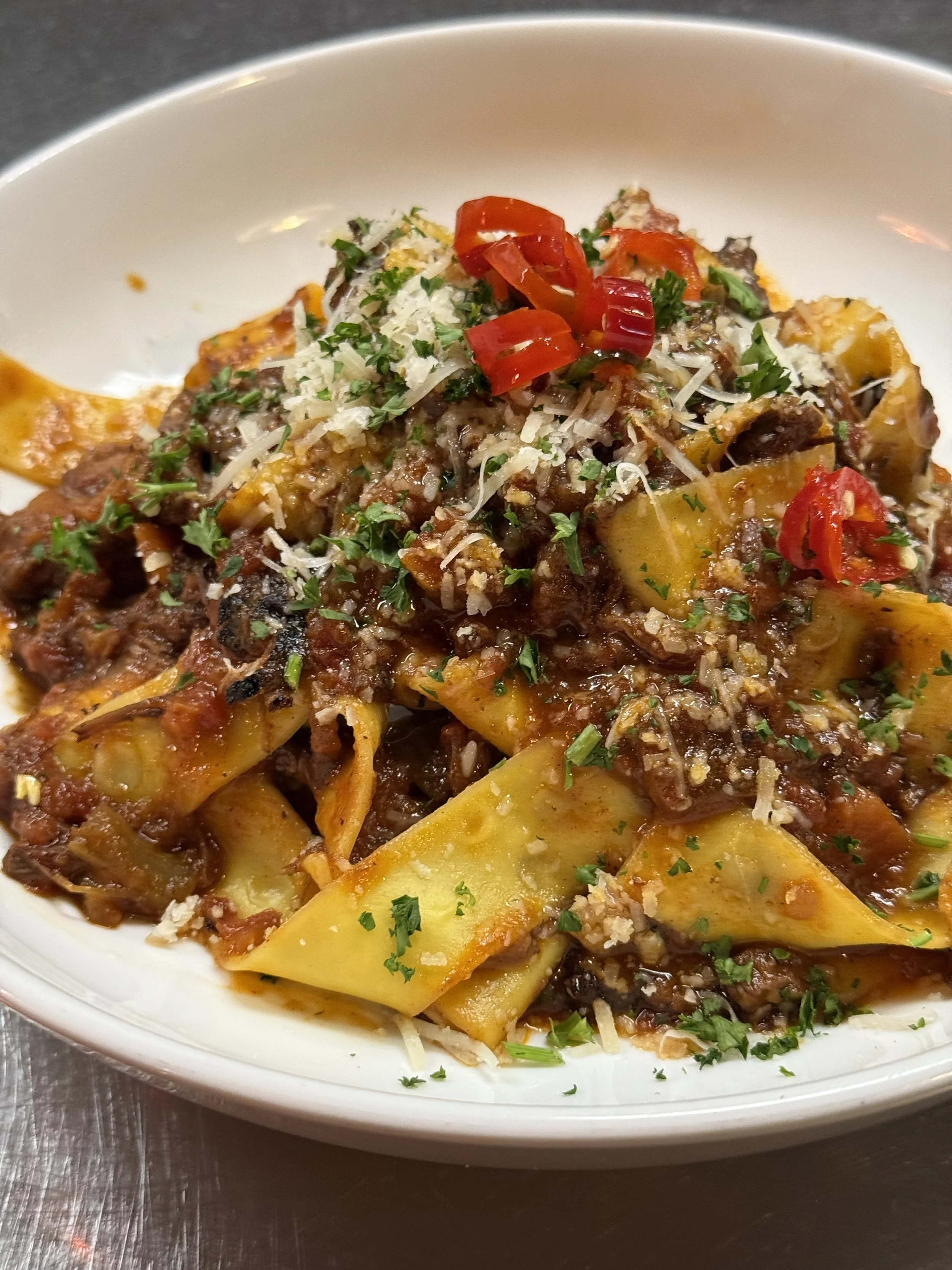 Close-up of a bowl of beef lasagna with cheese, chopped herbs, and chopped red and green chili peppers on top.