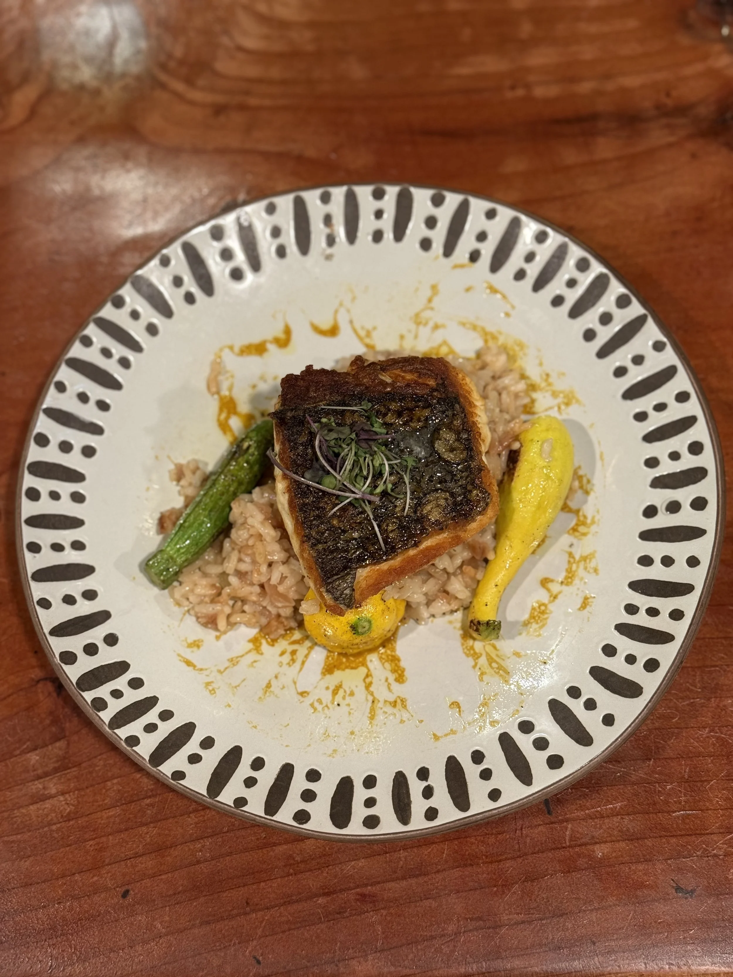 A plate of cooked rice topped with a grilled piece of fish, garnished with microgreens, accompanied by a green chili and a yellow chili on a decorative white plate with black patterns.