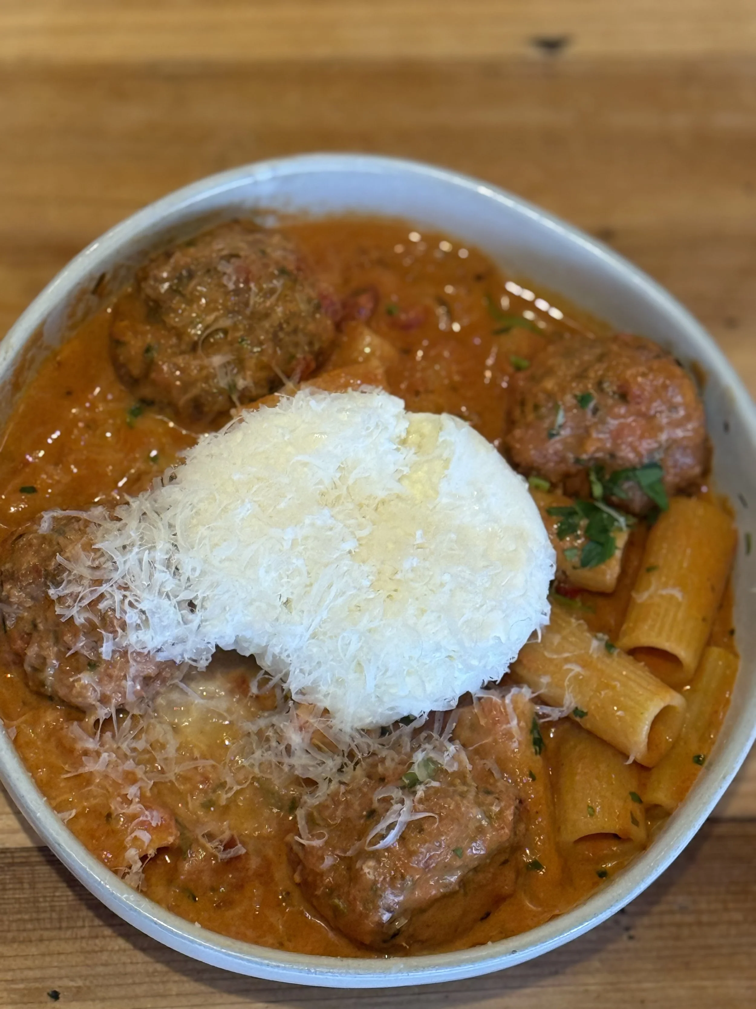 Bowl of Italian pasta with meatballs, topped with grated cheese and fresh herbs, served on a wooden table.