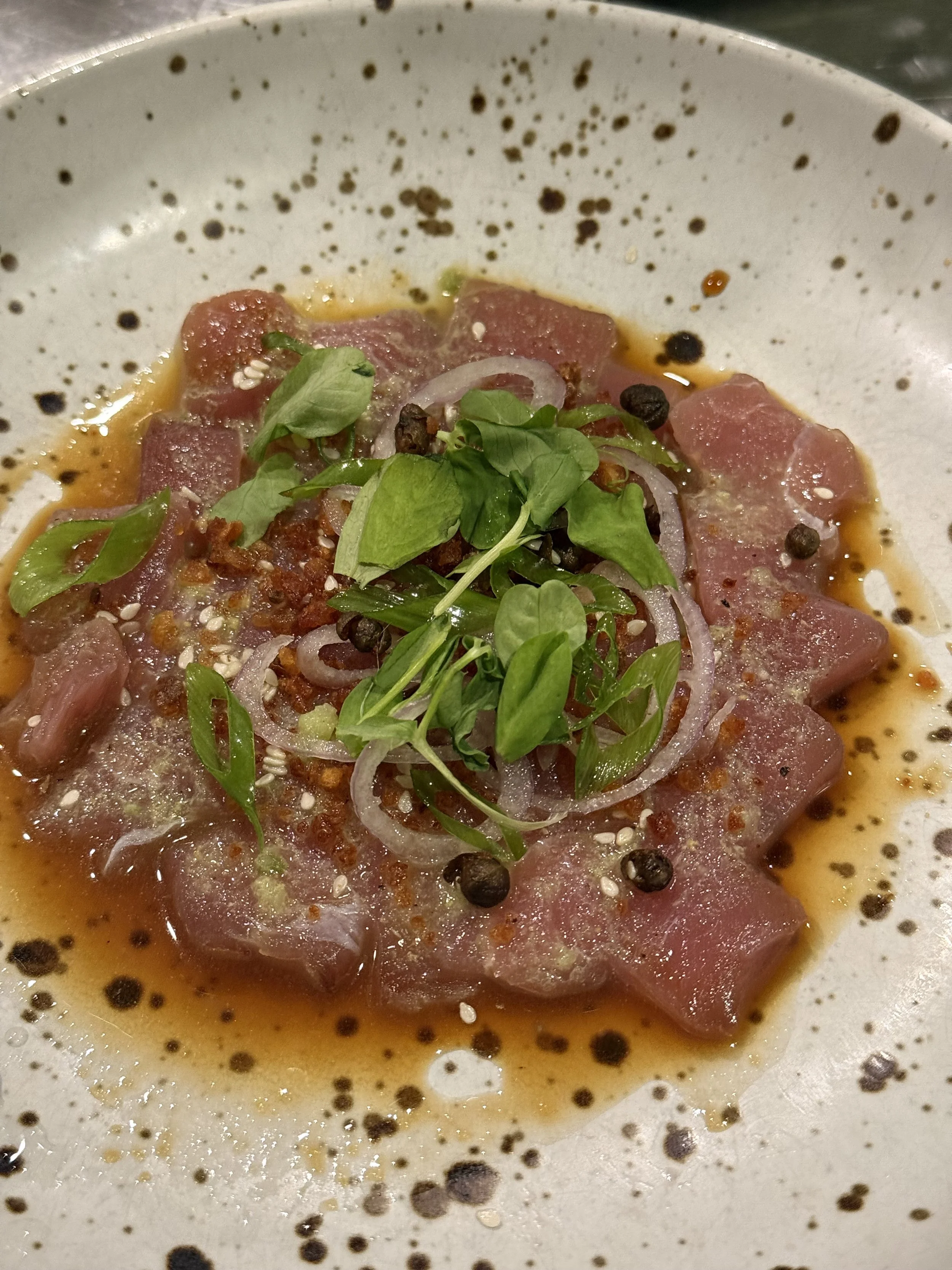 Sliced raw tuna tuna tuna with microgreens, onions, peppercorns, and sauce on a white speckled plate.