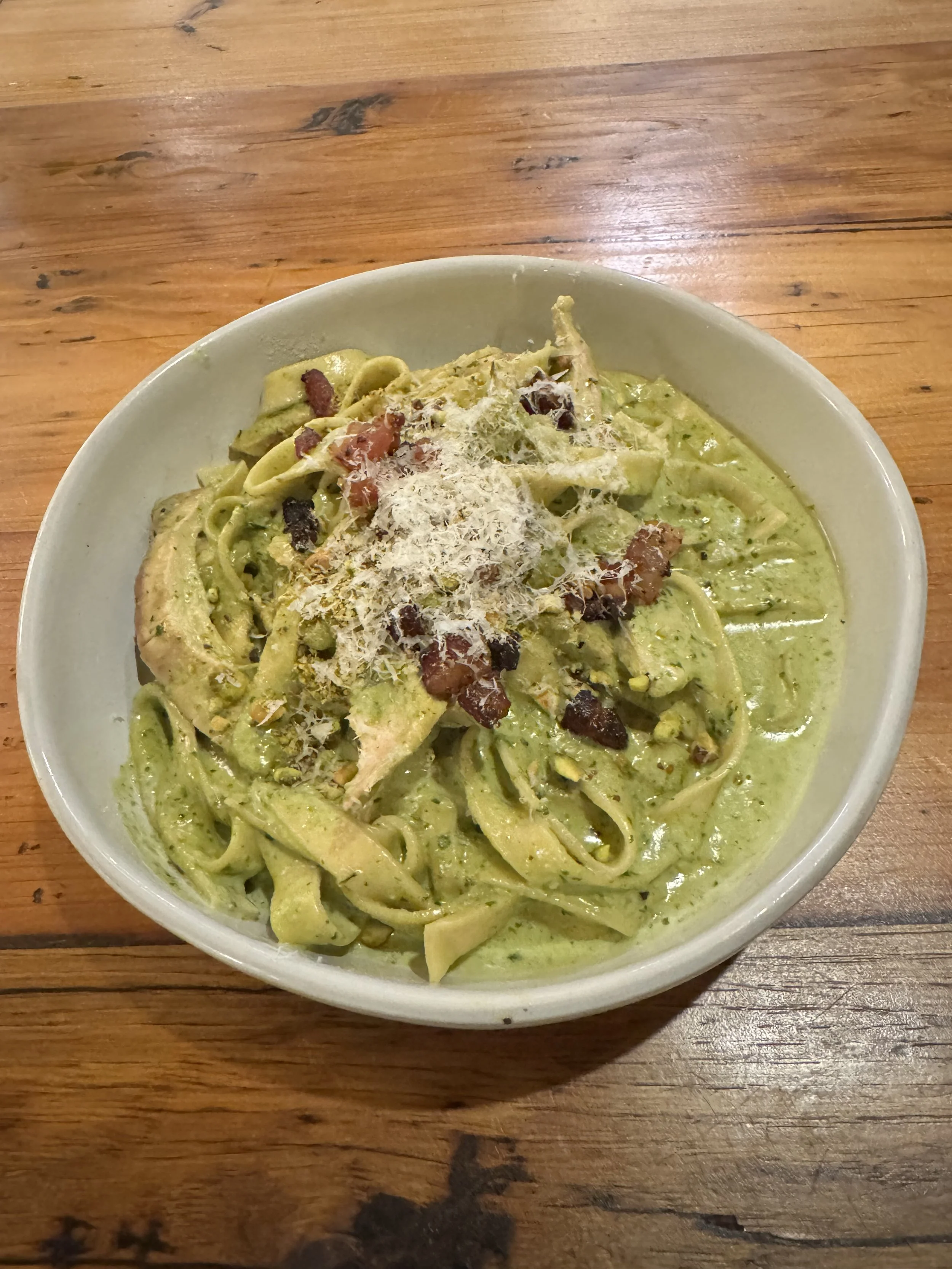A bowl of creamy green pasta topped with grated cheese, chopped nuts, and bits of bacon on a wooden table.