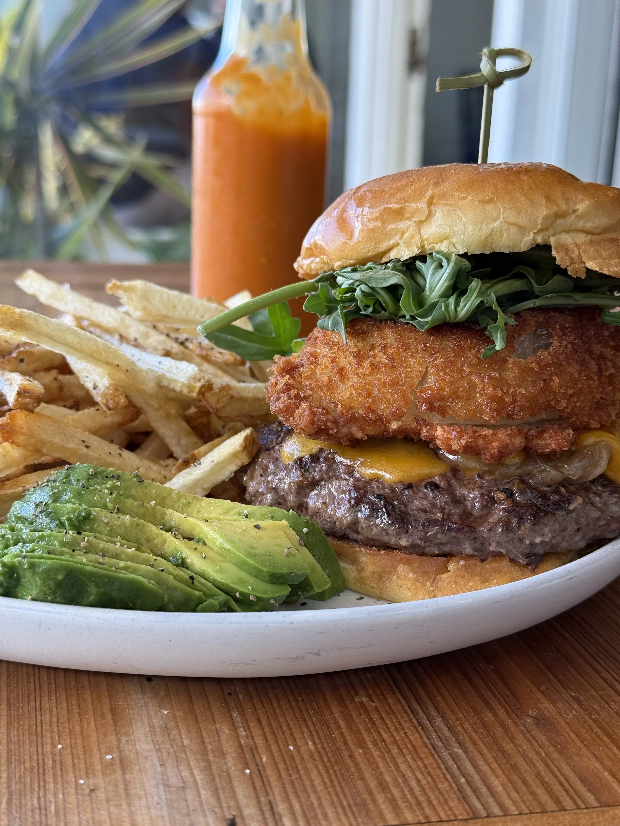 Close-up of a burger with fried chicken filet, lettuce, cheese, and bun, served with French fries and sliced avocado, with a bottle of orange sauce in the background.