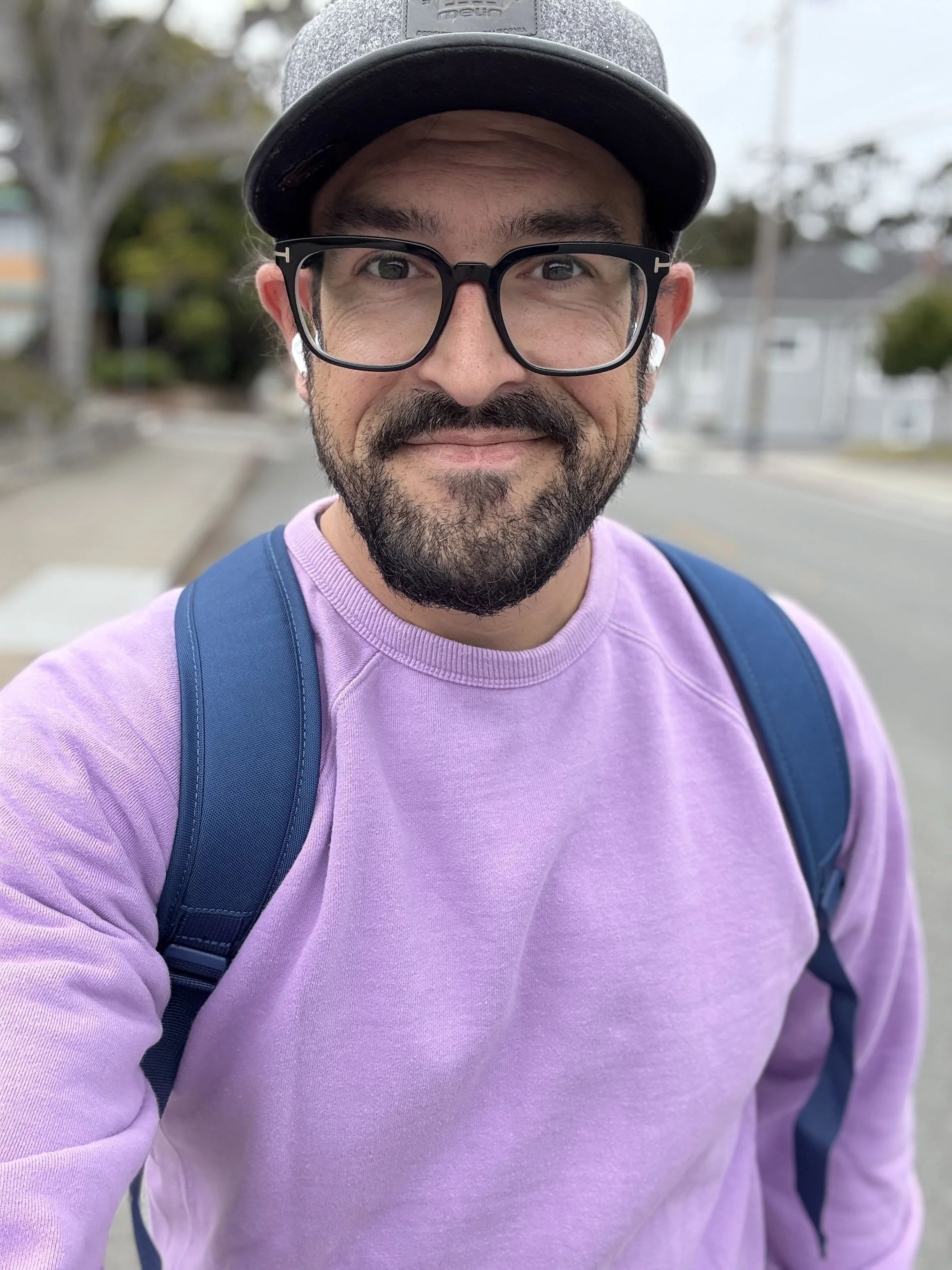A man taking a selfie outdoors during daytime, wearing glasses, a gray cap, a purple sweatshirt, and a blue backpack.