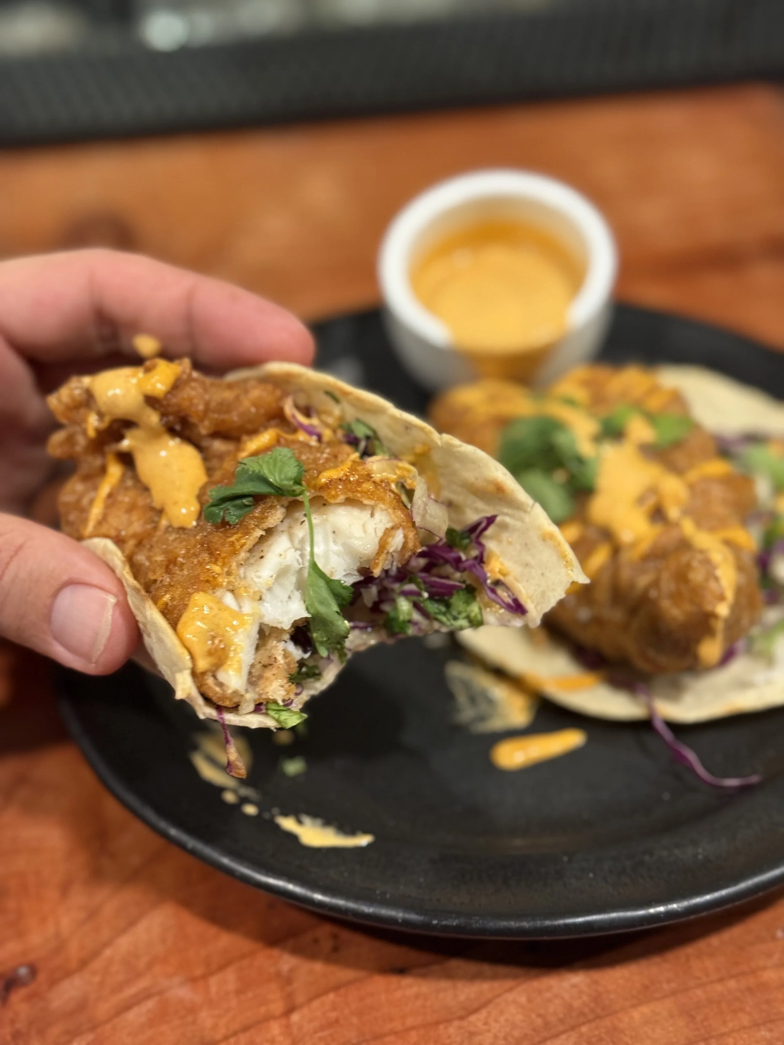 Close-up of a hand holding a partially eaten taco with fried chicken, shredded lettuce, cilantro, and cheese, on a black plate with more tacos and a cup of orange-colored sauce in the background.