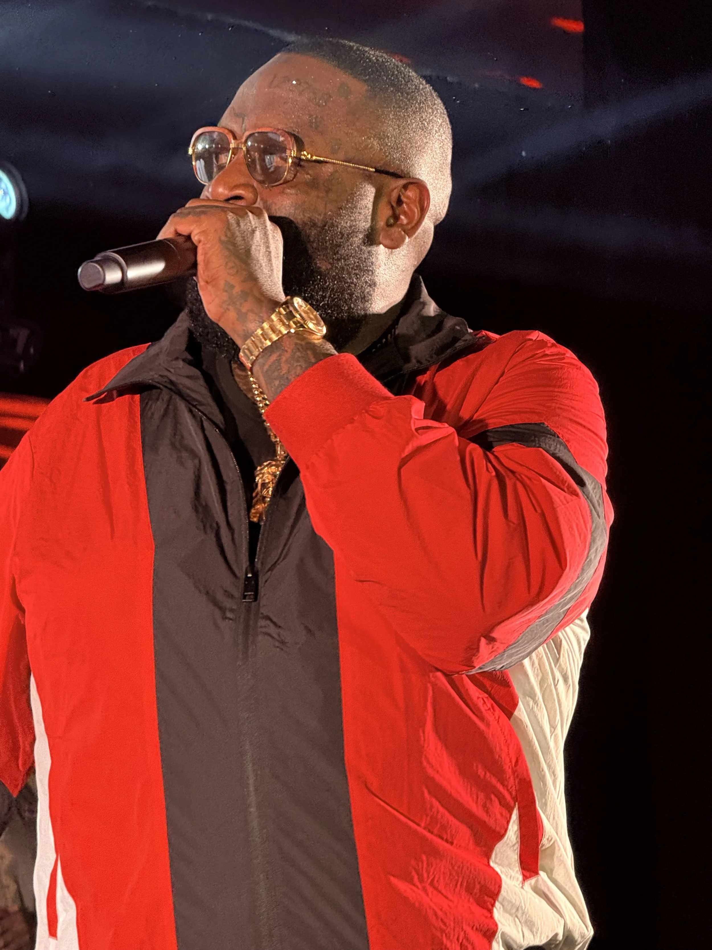 A man with sunglasses and tattoos on his hand is holding a microphone and singing or speaking. He is wearing a red and black jacket and has a gold watch and jewelry. The background is dark with stage lighting.