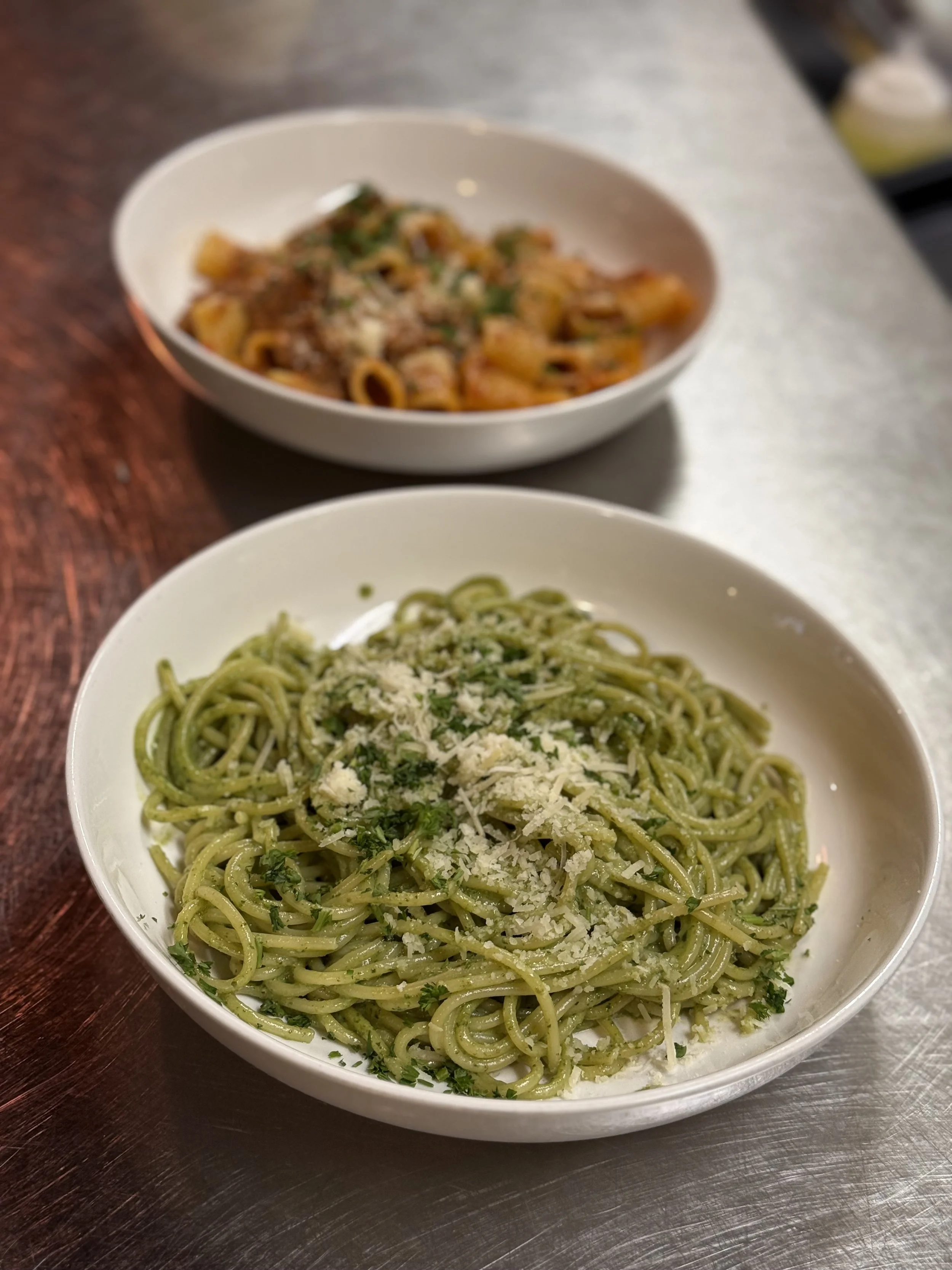 A bowl of green pesto pasta topped with grated cheese and parsley, with another bowl of tomato-based pasta in the background.