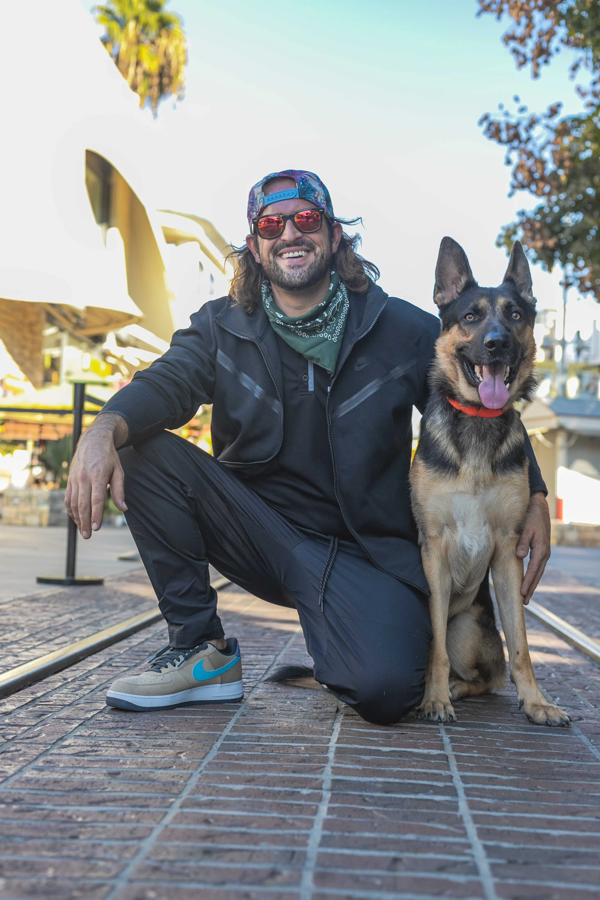 A man squatting on a brick sidewalk with his arm around a sitting German Shepherd dog. The man is smiling, wearing sunglasses, a backward baseball cap, a black jacket, black pants, grey and blue Nike sneakers, and a green bandana. The dog has its tongue out, wearing a red collar. In the background, there are trees, a canopy, and a building, under a clear blue sky.