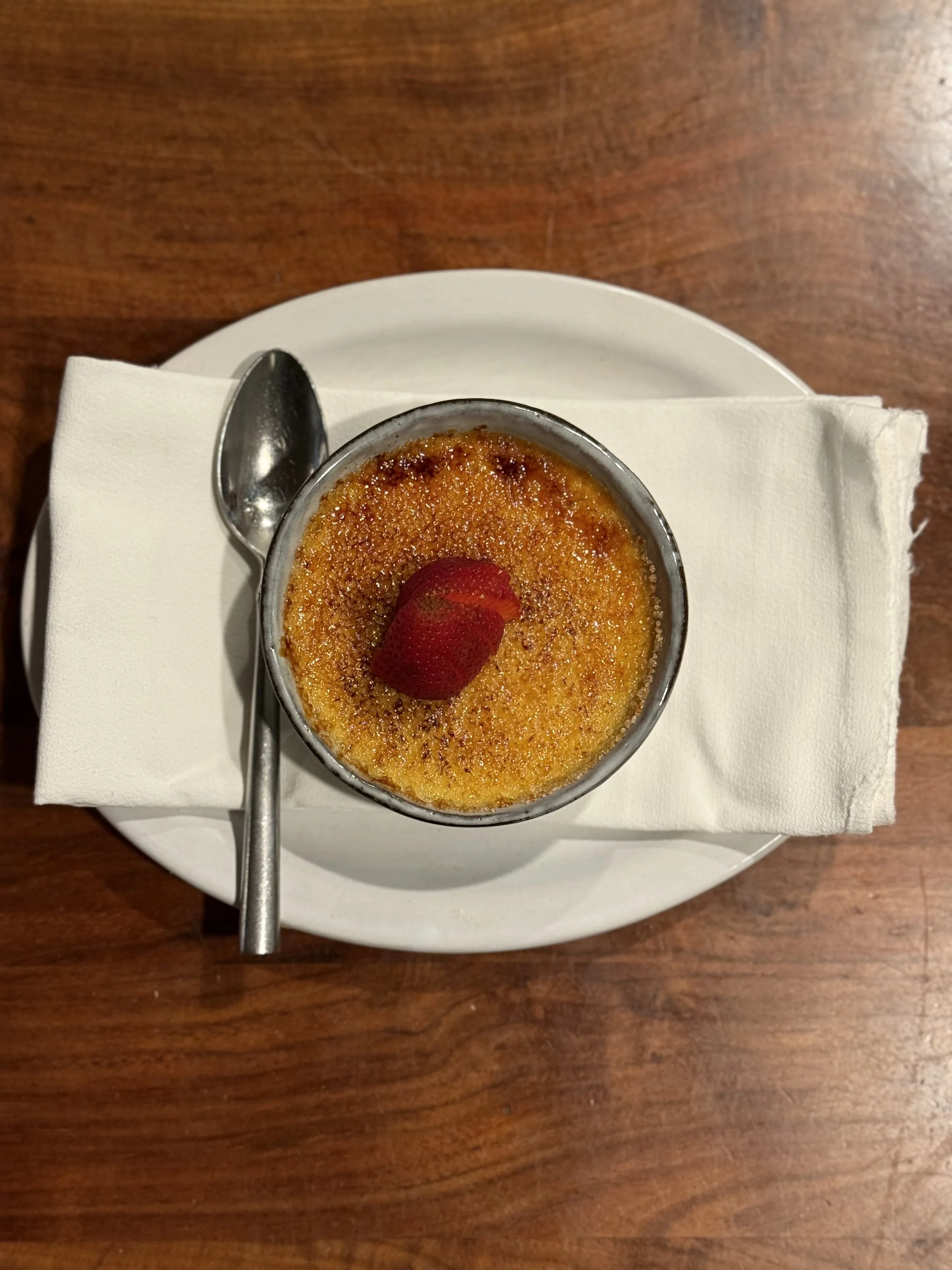 A serving of crème brûlée garnished with a strawberry on top, placed on a white plate with a spoon on a napkin, on a wooden table.