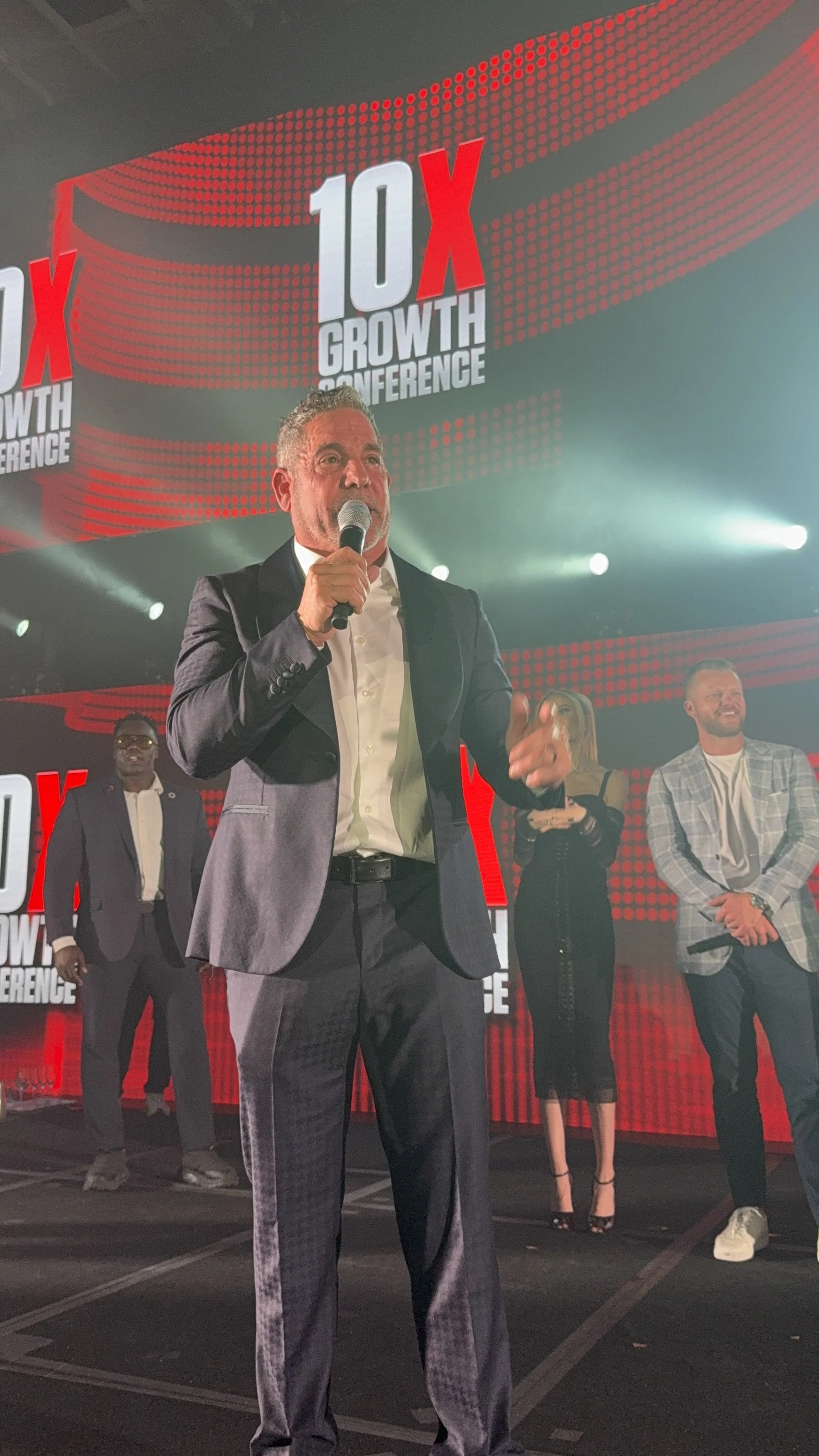 A man in a suit speaking into a microphone on stage during the 10X Growth Conference, with others standing behind him and a large screen displaying the event's logo.