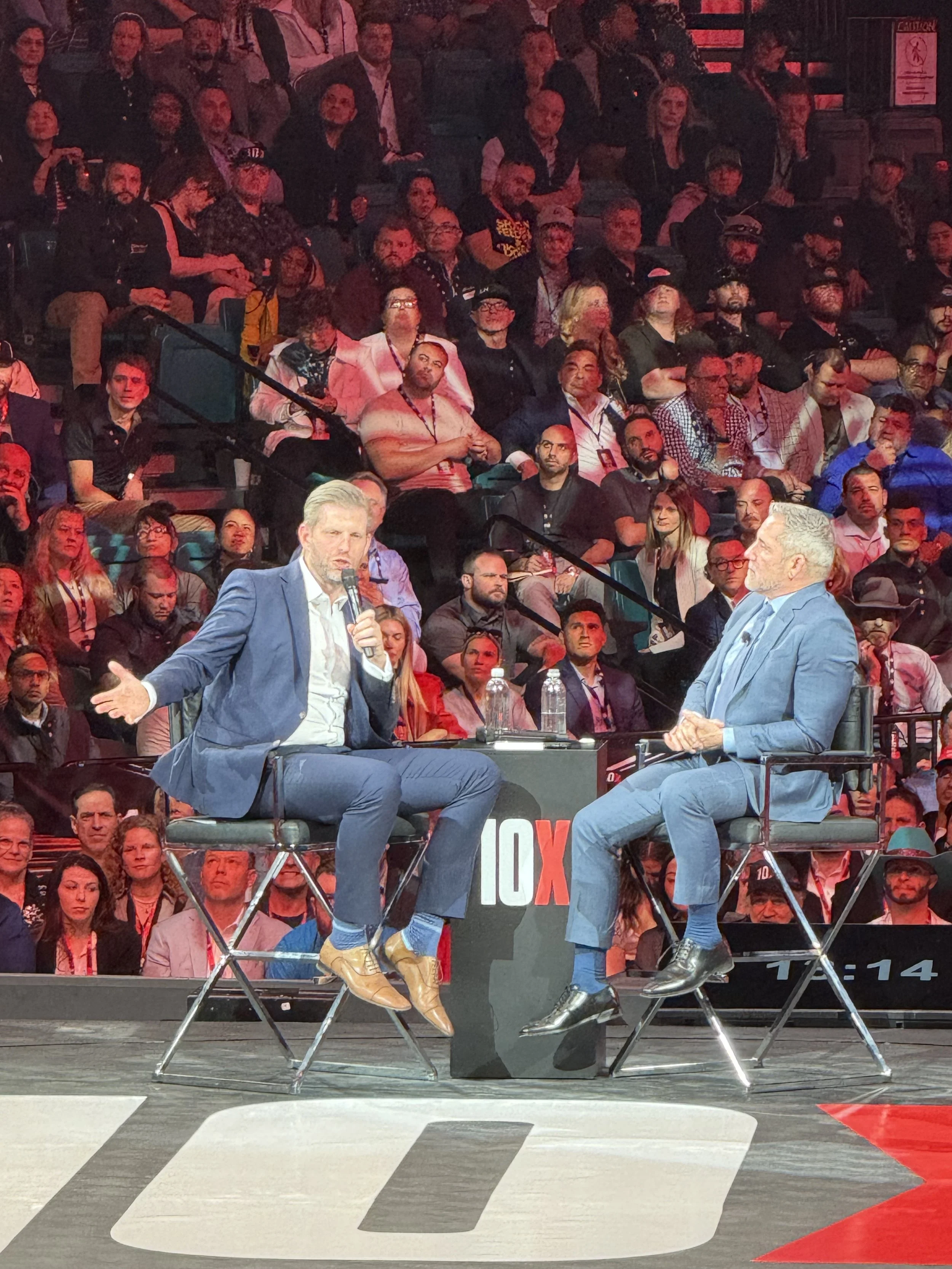 Two men in suits are seated on stage, engaged in a conversation with one man holding a microphone. They are facing each other, with a large audience watching in the background during a televised event, indicated by the TEDx and TV screen branding.