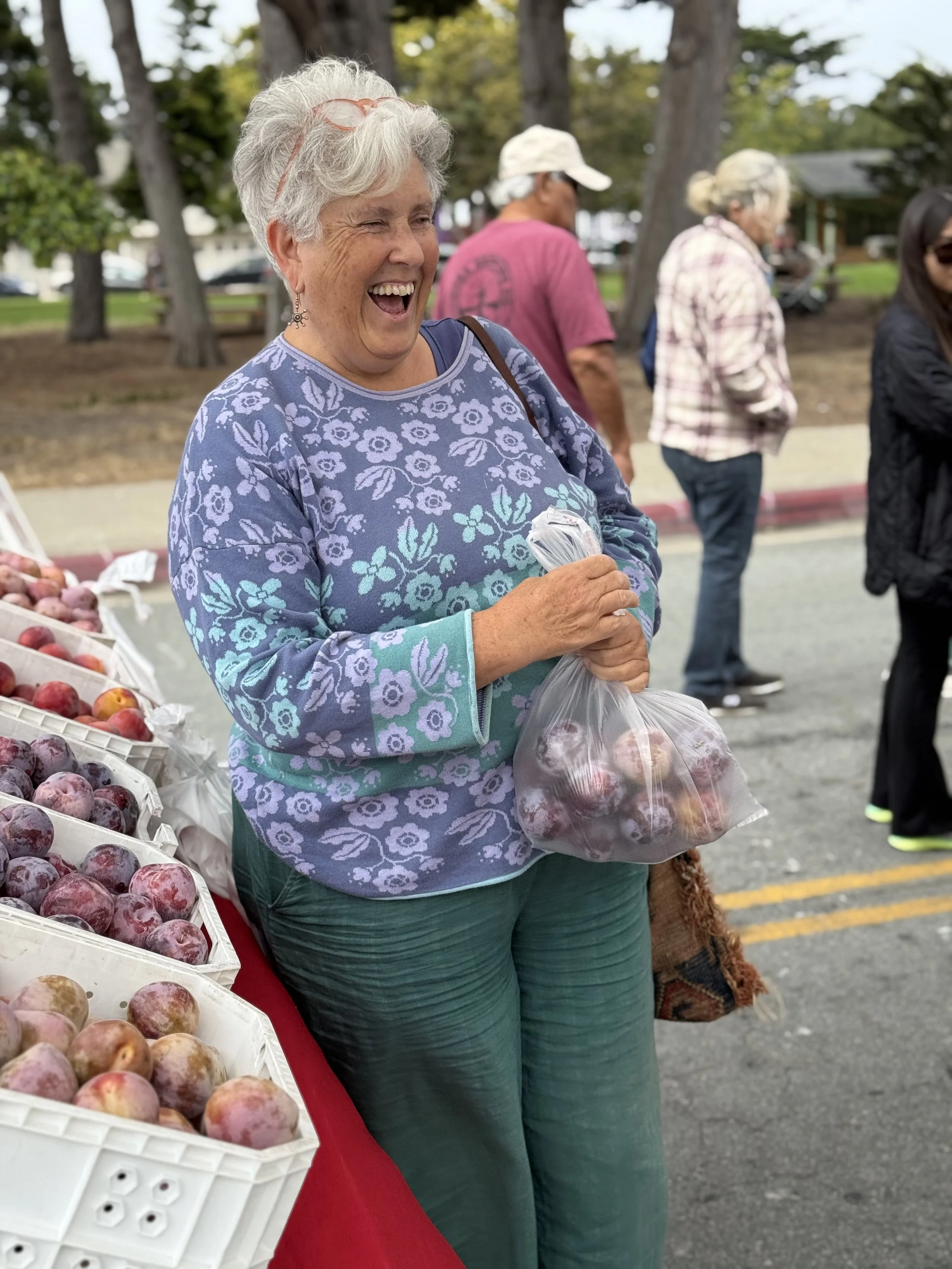 A cheerful older woman with short gray hair and glasses on her head, holding a plastic bag of apples at an outdoor farmers market. She is wearing a blue floral sweater and green pants, smiling as she shops.