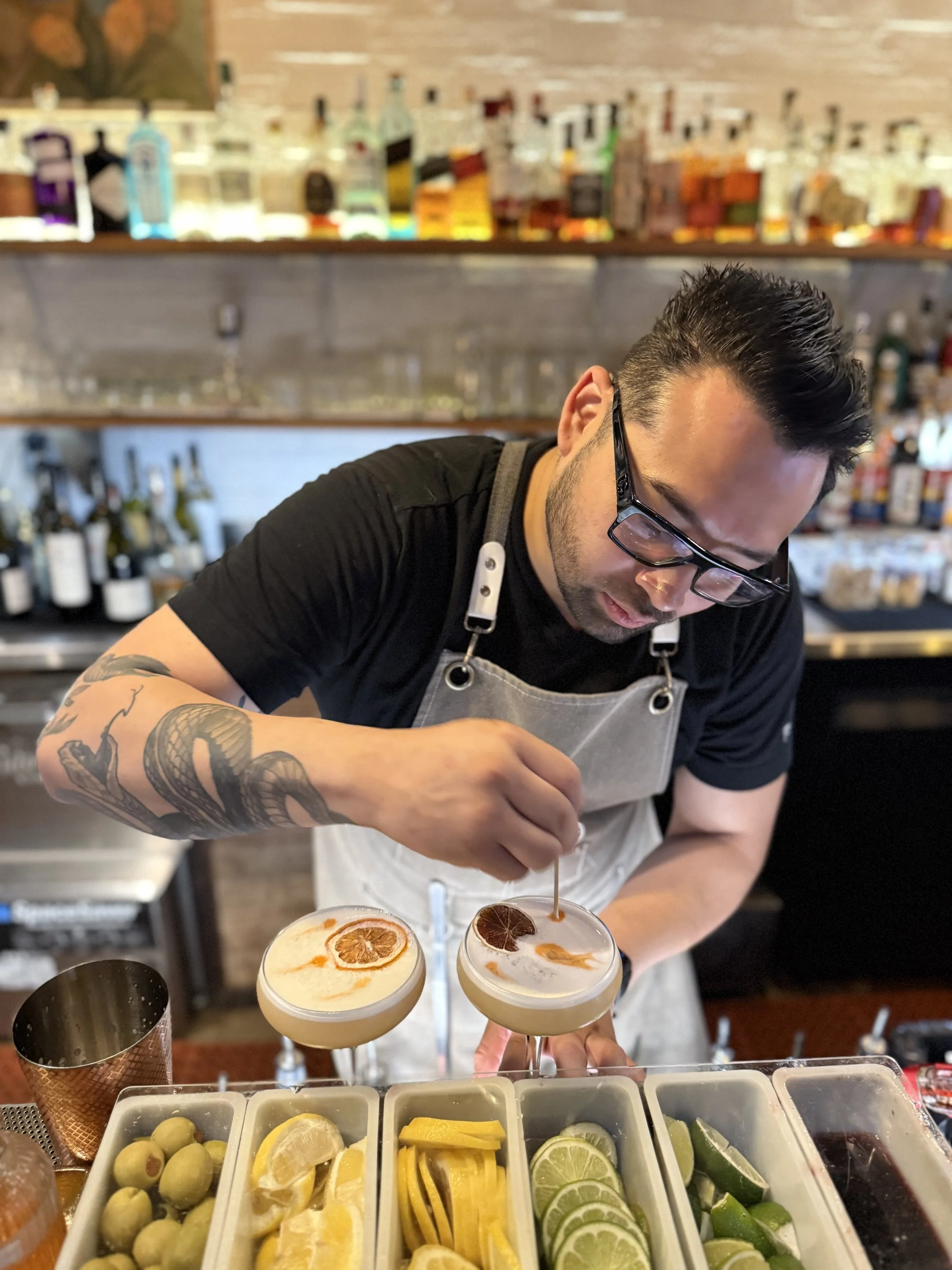 A bartender with glasses and tattoos on his arm preparing cocktails at a bar with bottles and fruit garnishes in the background.