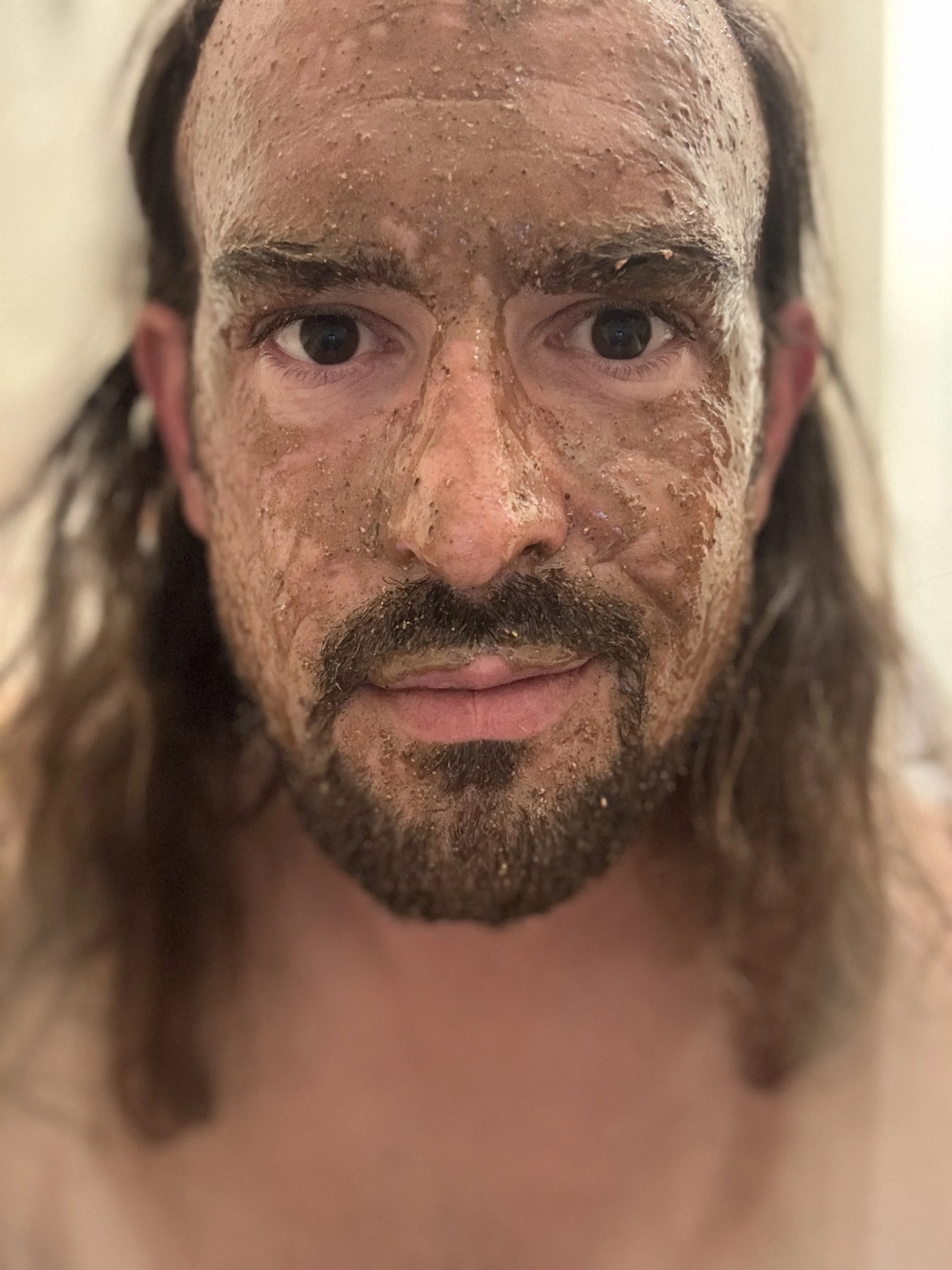 A man with long hair and a beard has a facial mask applied, which appears to be a mud or clay mask, and it covers his face from forehead to chin.