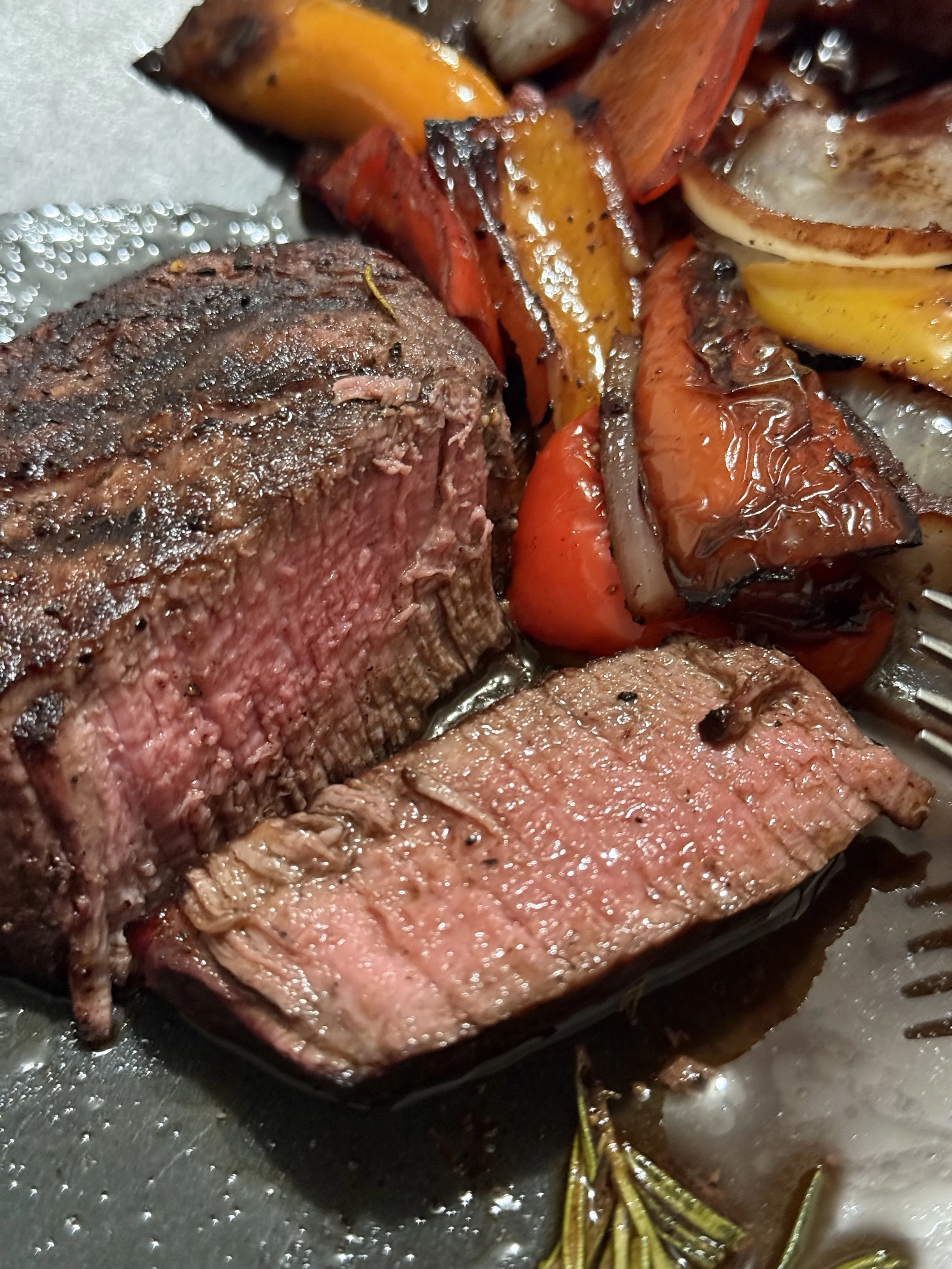 Close-up of a cooked steak with a pink center next to grilled vegetables including tomatoes, bell peppers, onions, and zucchini, with a sprig of rosemary.