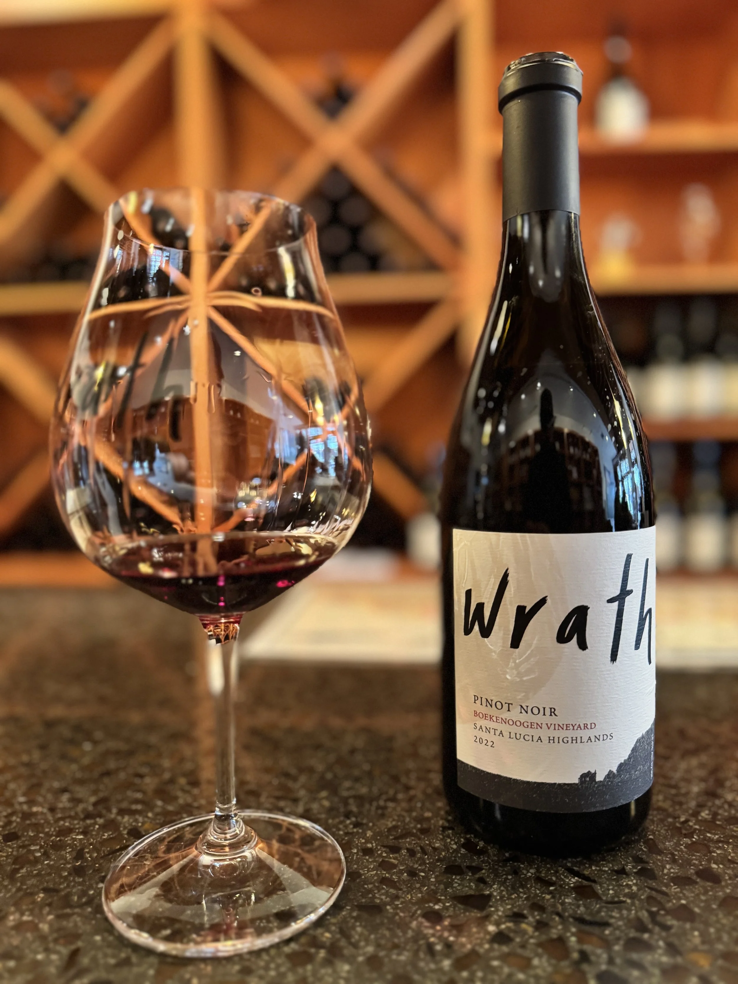 A glass of red wine and a bottle of Wrath Pinot Noir wine on a kitchen counter with a wine rack in the background.