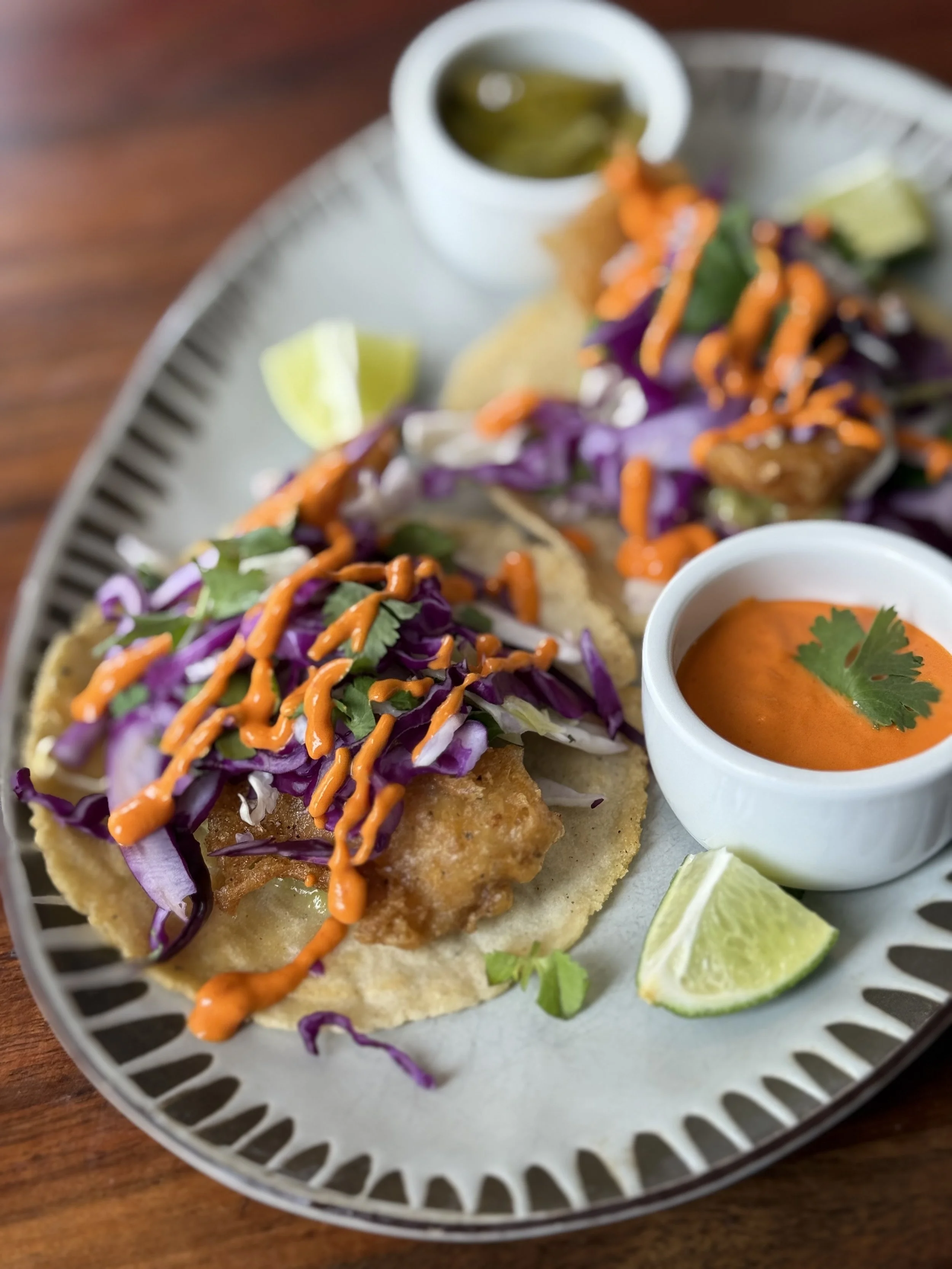 Close-up of fish tacos topped with shredded purple cabbage, cilantro, and orange sauce, served with lime wedges, creamy orange sauce with cilantro, and a small cup of green salsa on a silver tray.