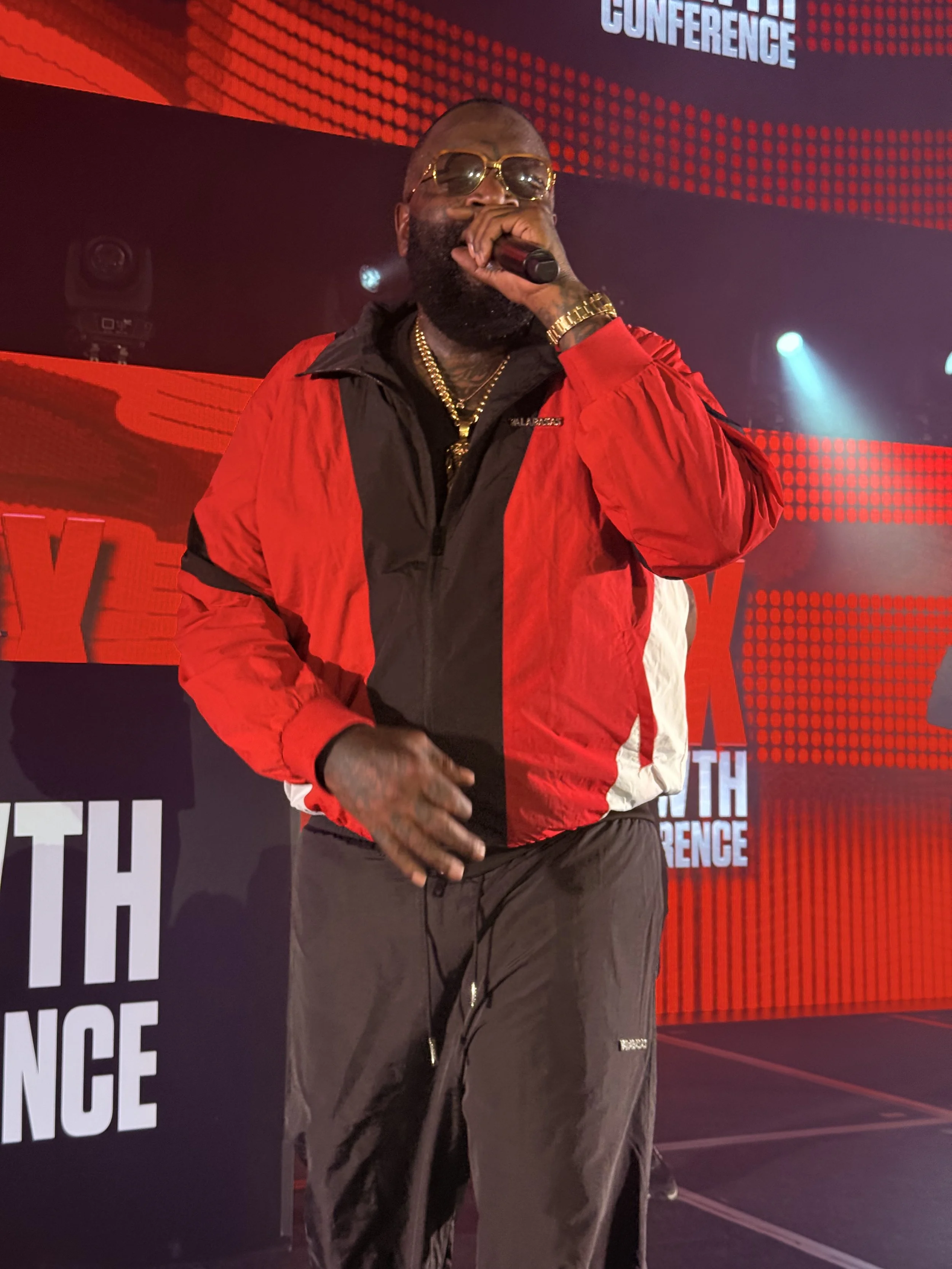 A man performing on stage with a microphone, wearing sunglasses, a red and black jacket, gold chains, and a gold watch, with a background displaying red and black graphics and the words 'Youth Conference'.