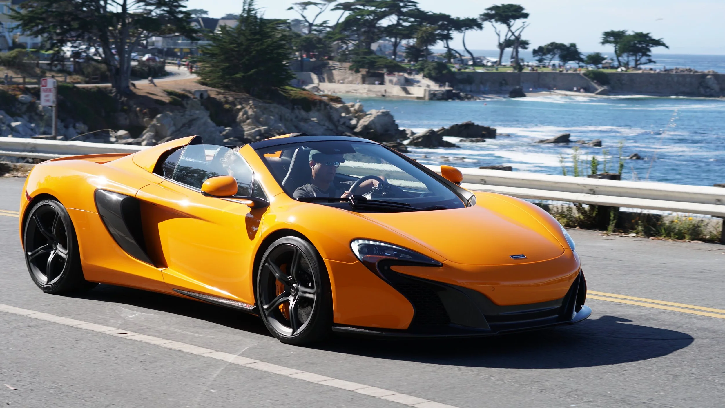 An orange sports car driving along a coastal road with ocean and rocky shoreline in the background.