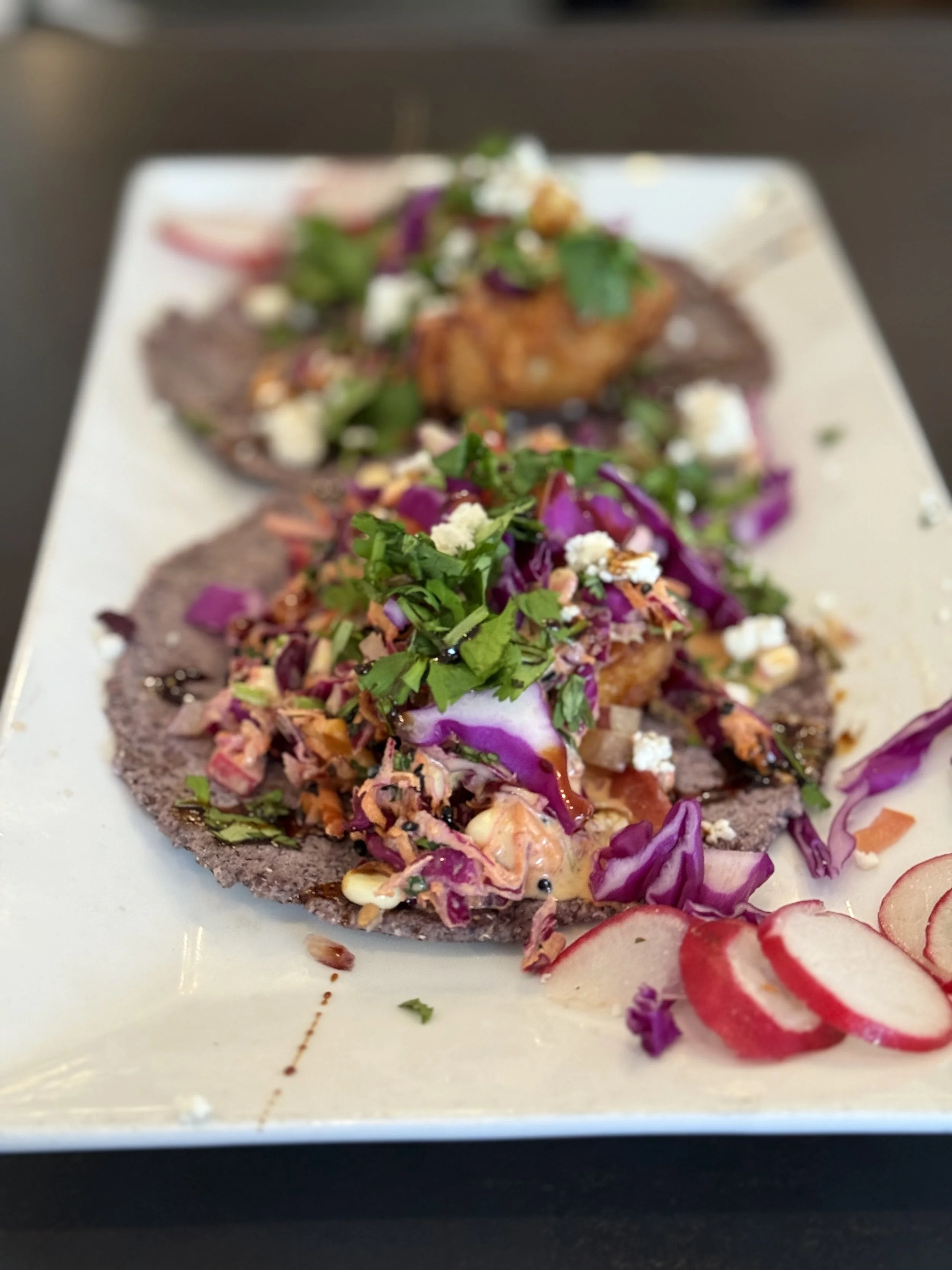 Tacos with purple corn tortillas filled with coleslaw, greens, and toppings, garnished with sliced radishes.