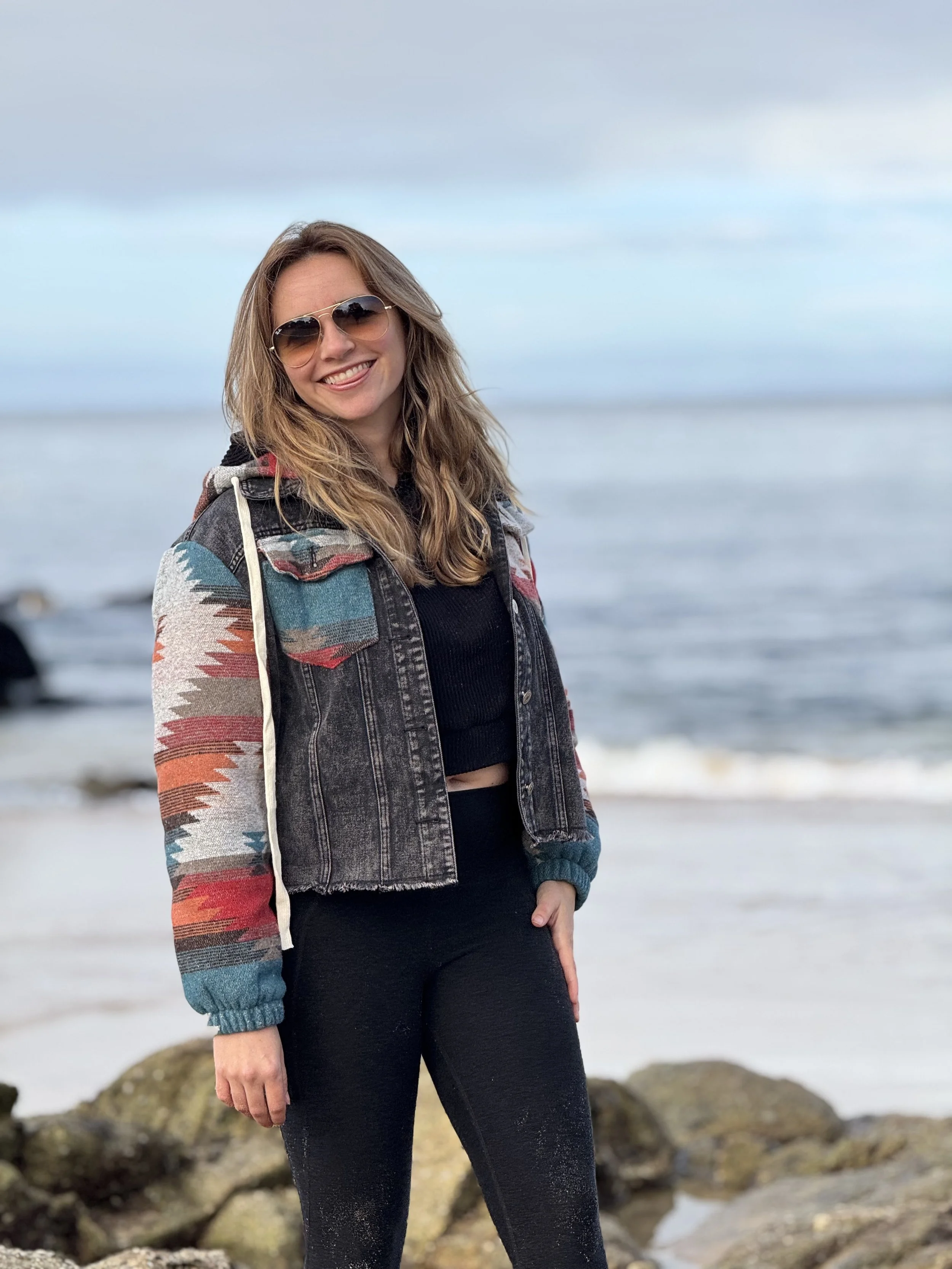 Smiling woman in sunglasses and a colorful patterned jacket standing on rocks at the beach with ocean and cloudy sky in the background.