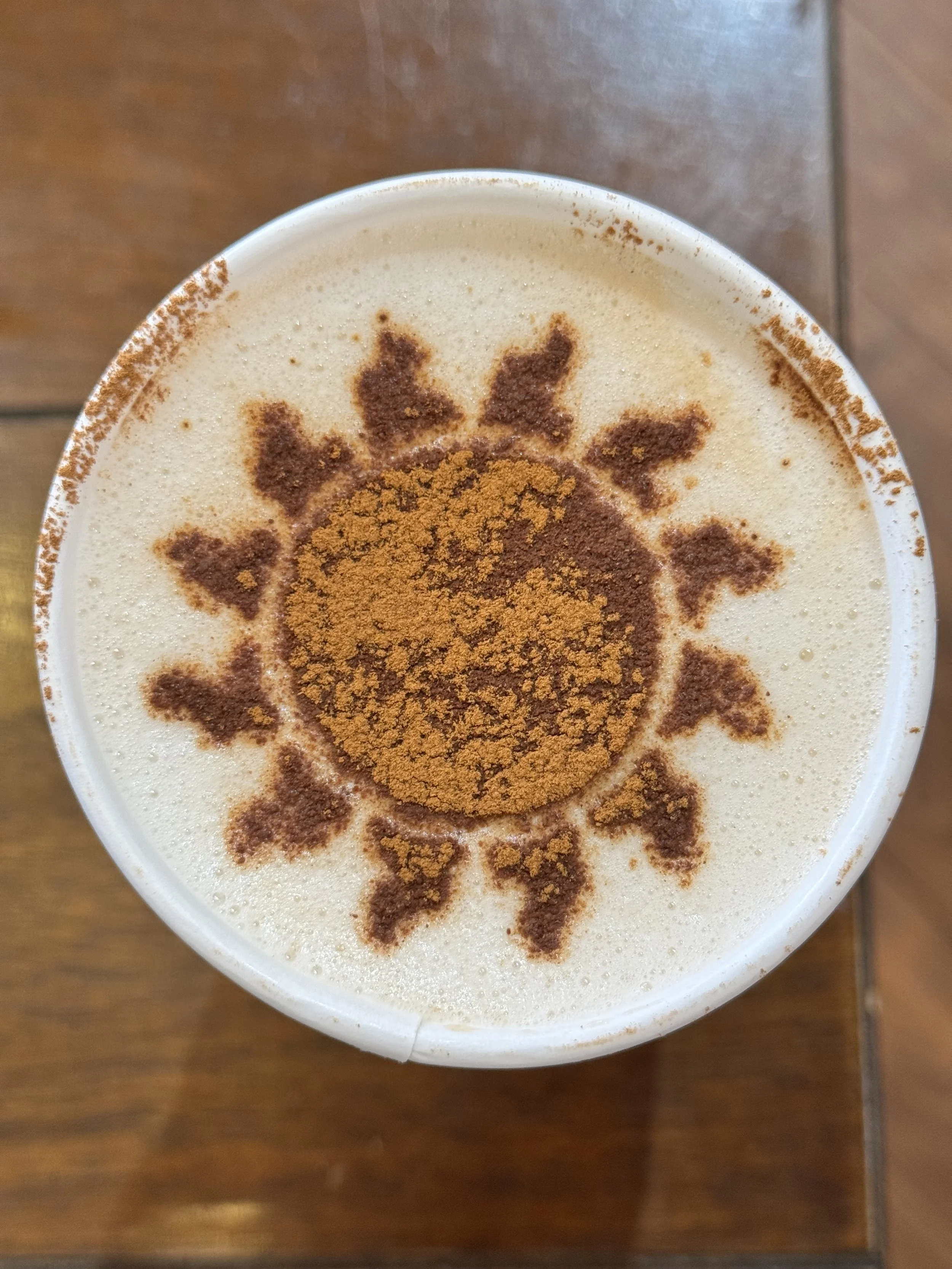 A top view of a cup of frothy coffee with a sun pattern decorated on top using cinnamon or cocoa powder.