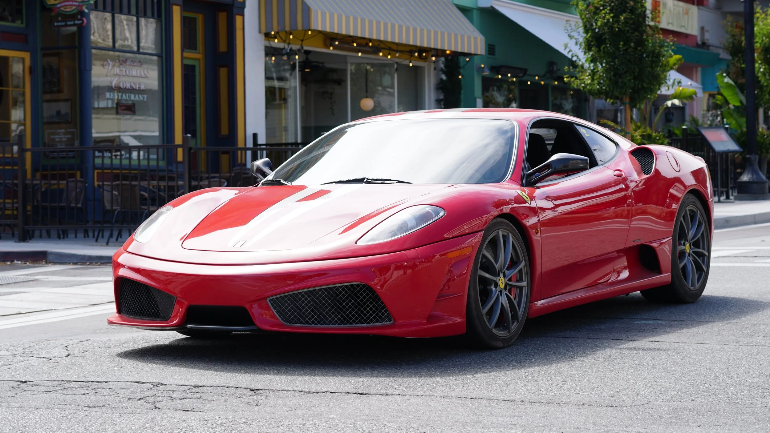 Red sports car with white racing stripe parked on street in front of restaurant with outdoor seating area.