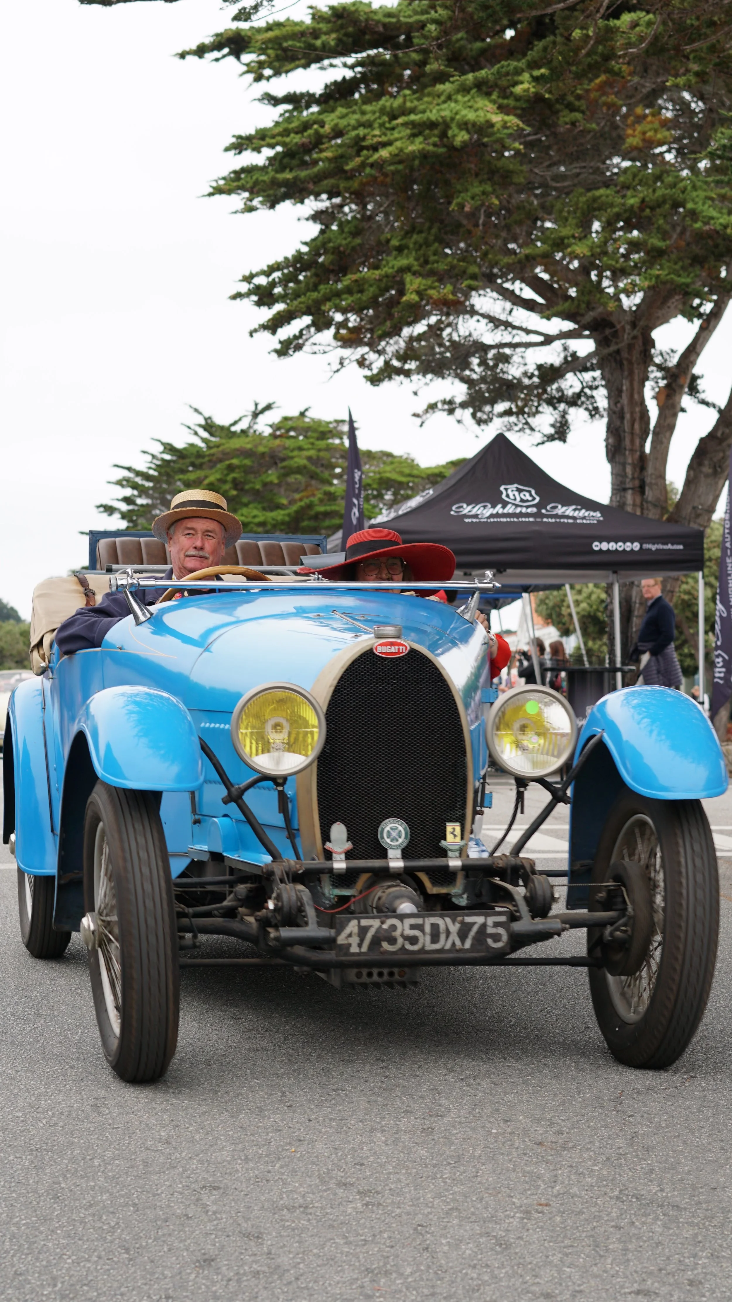 A vintage blue Bugatti car with two people inside, wearing hats, at an outdoor event. The background includes a black tent and trees.