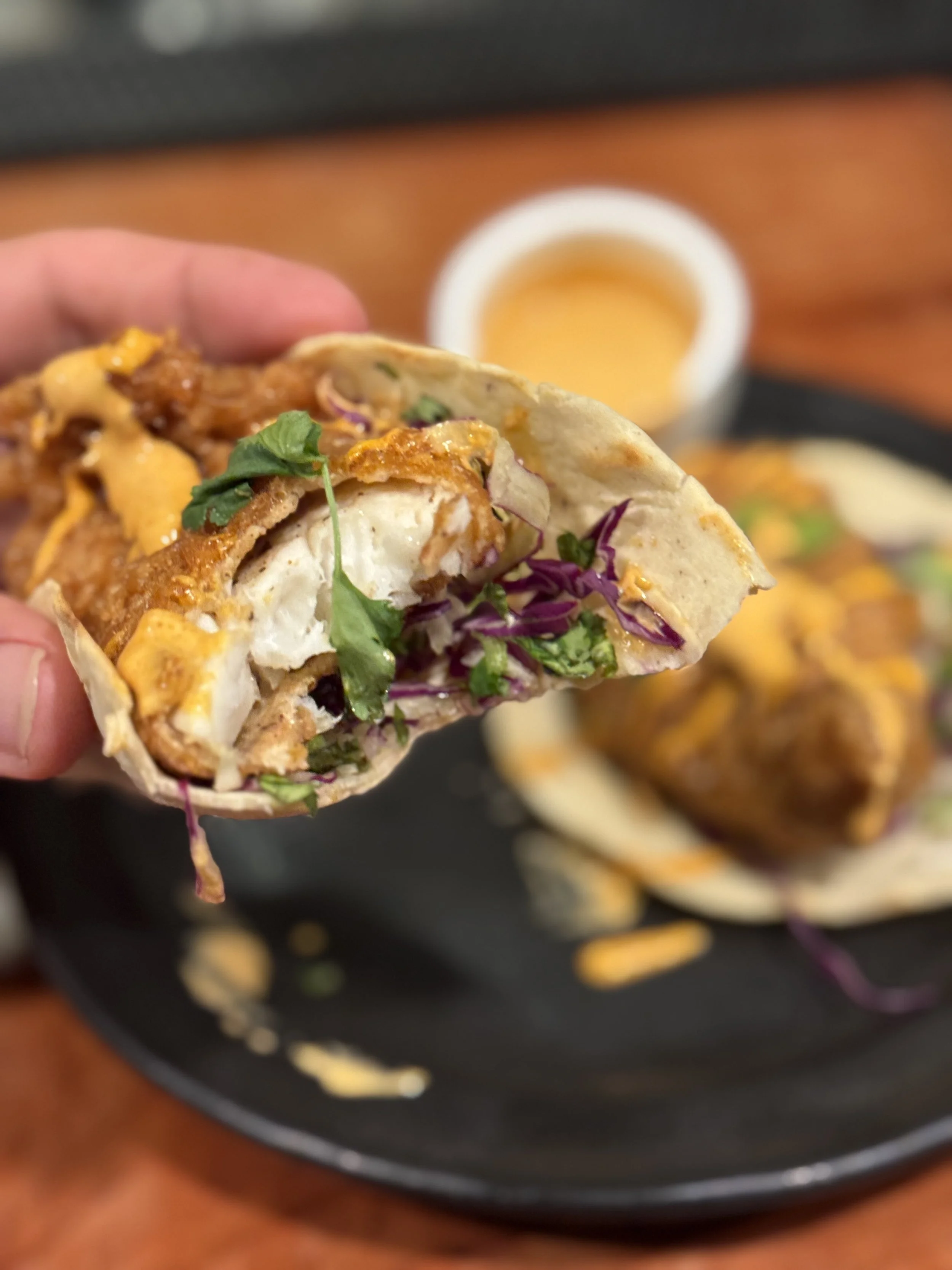 Close-up of a taco with chicken, cheese, and vegetables, held in hand over a platter with more tacos and a small container of sauce in the background.