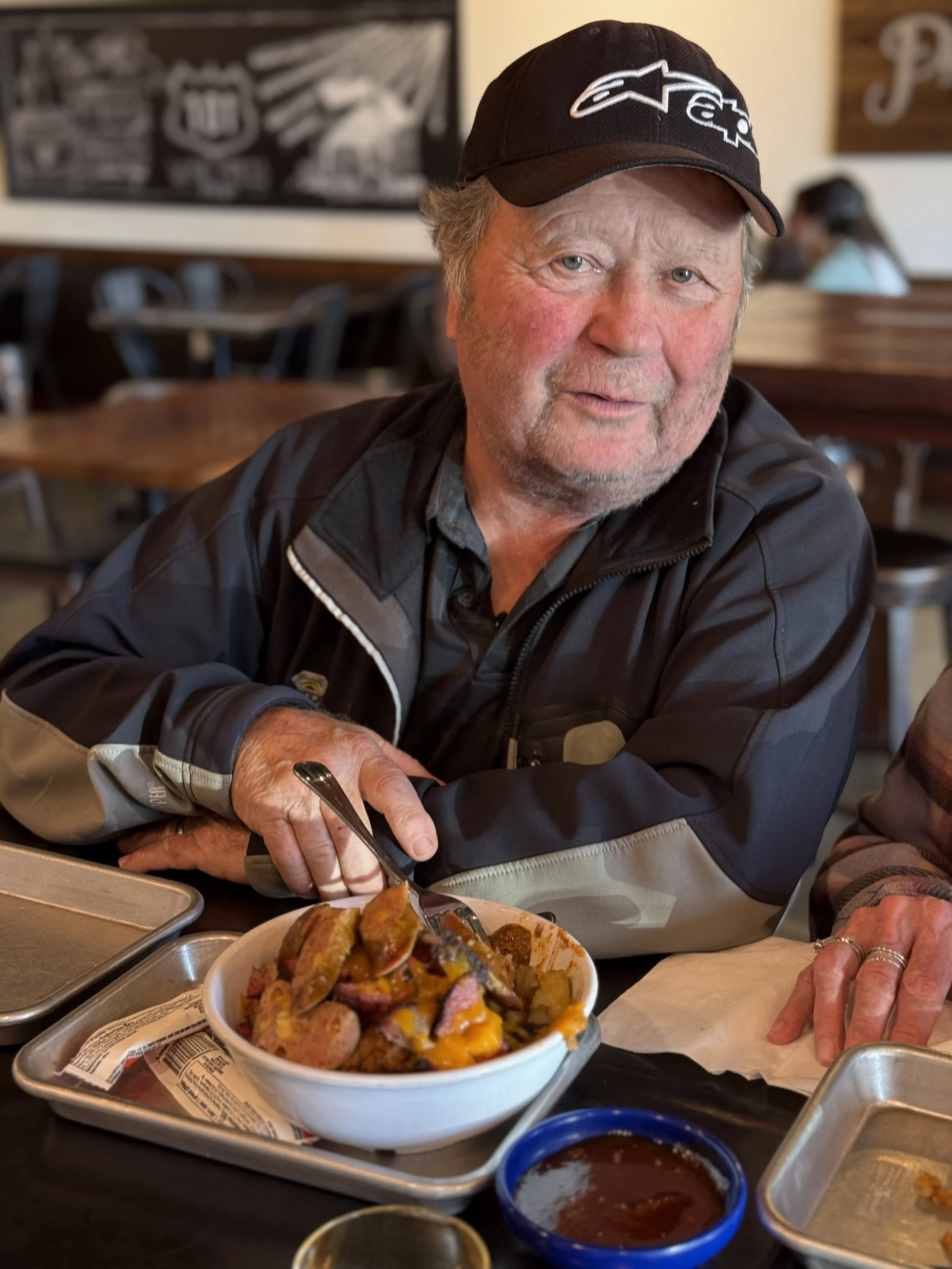 An older man with a beard and a black baseball cap sitting at a restaurant table with a bowl of food, holding a fork, and smiling. In the background, other people are dining.