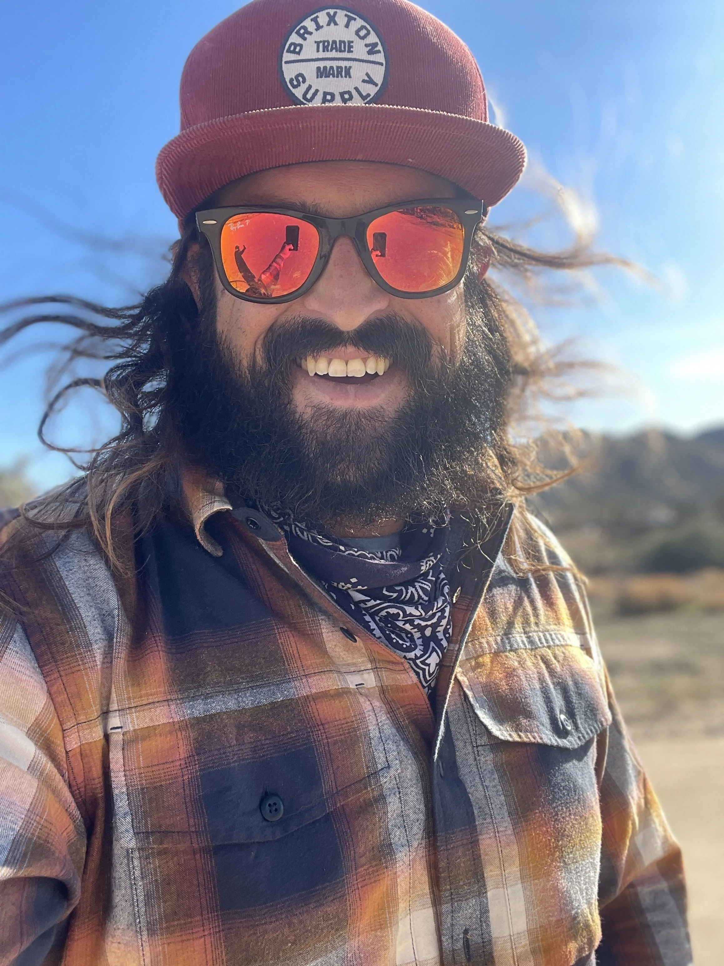 Man wearing a red cap with a white and blue logo, reflective sunglasses, a plaid shirt, and a bandana, smiling outdoors on a sunny day.