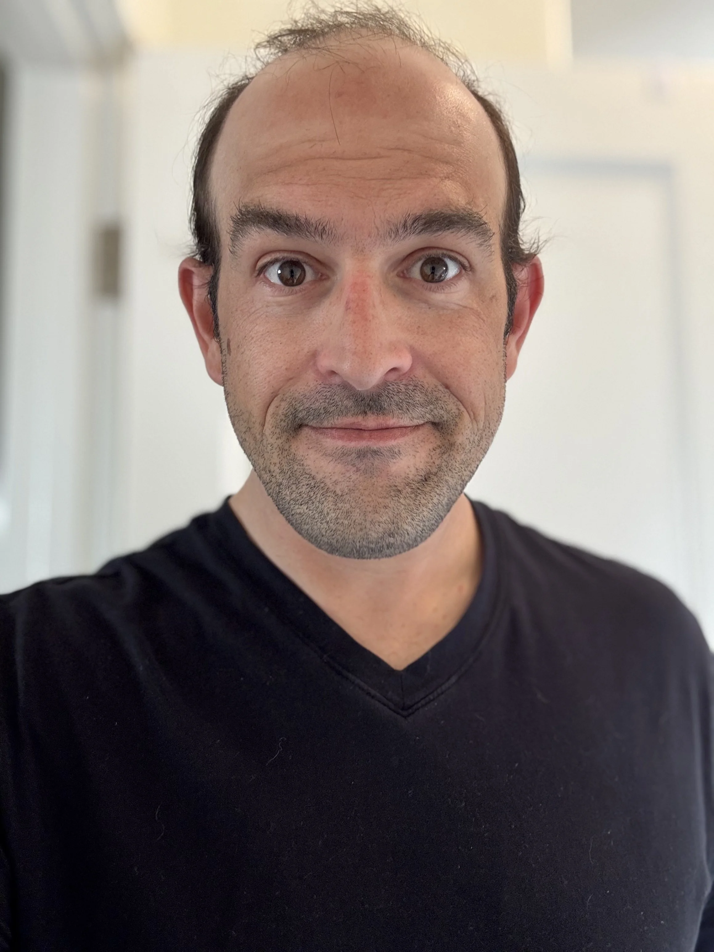 A man taking a selfie indoors, wearing a black shirt, smiling slightly with a neutral background.
