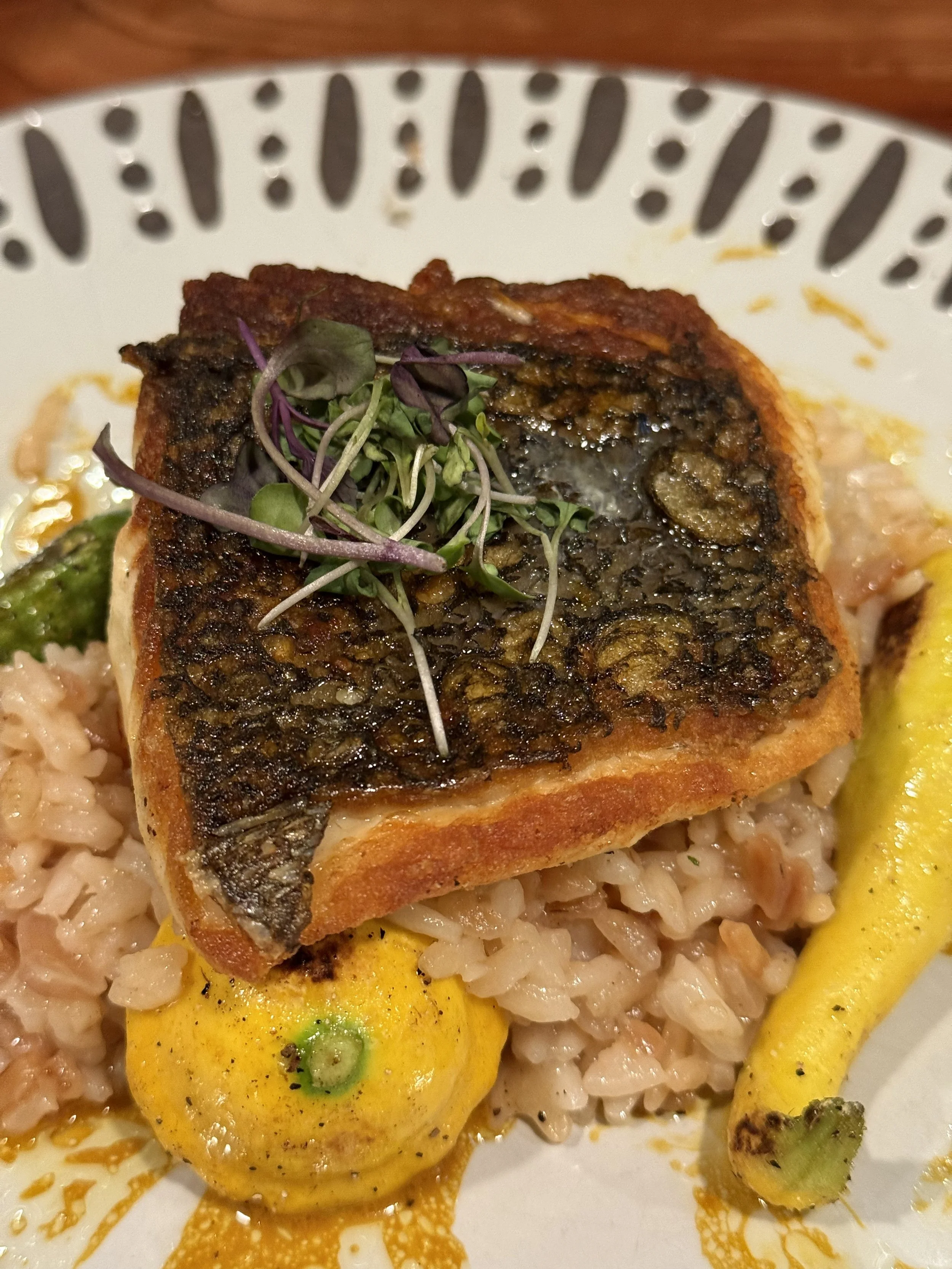 A plated dish featuring a cooked fish fillet with crispy skin, garnished with microgreens, served over rice with a yellow squash and a yellow chili pepper on the side.