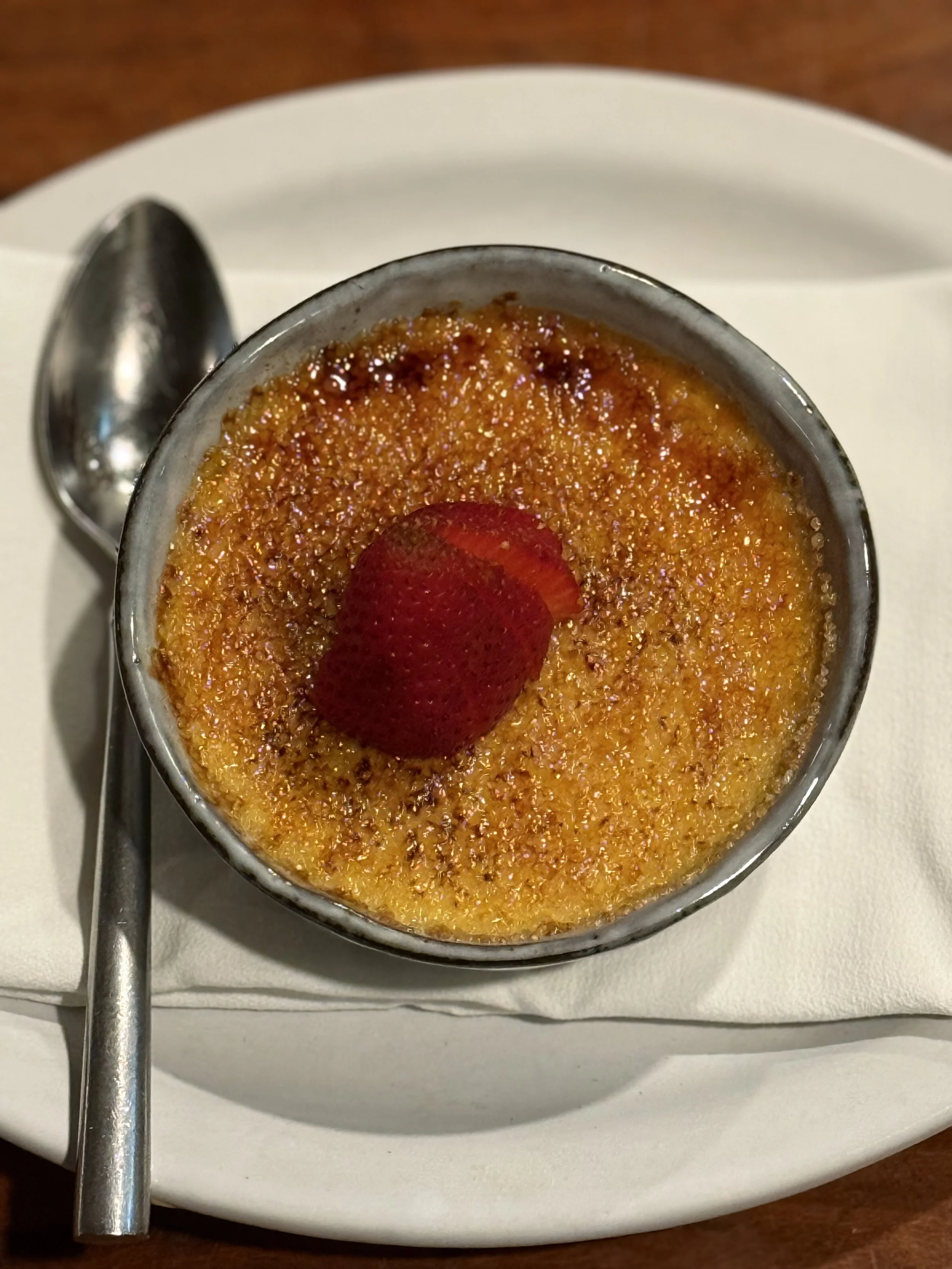 Crème brûlée with caramelized sugar topping, garnished with a strawberry half on top, served on a white plate with a spoon on the side.