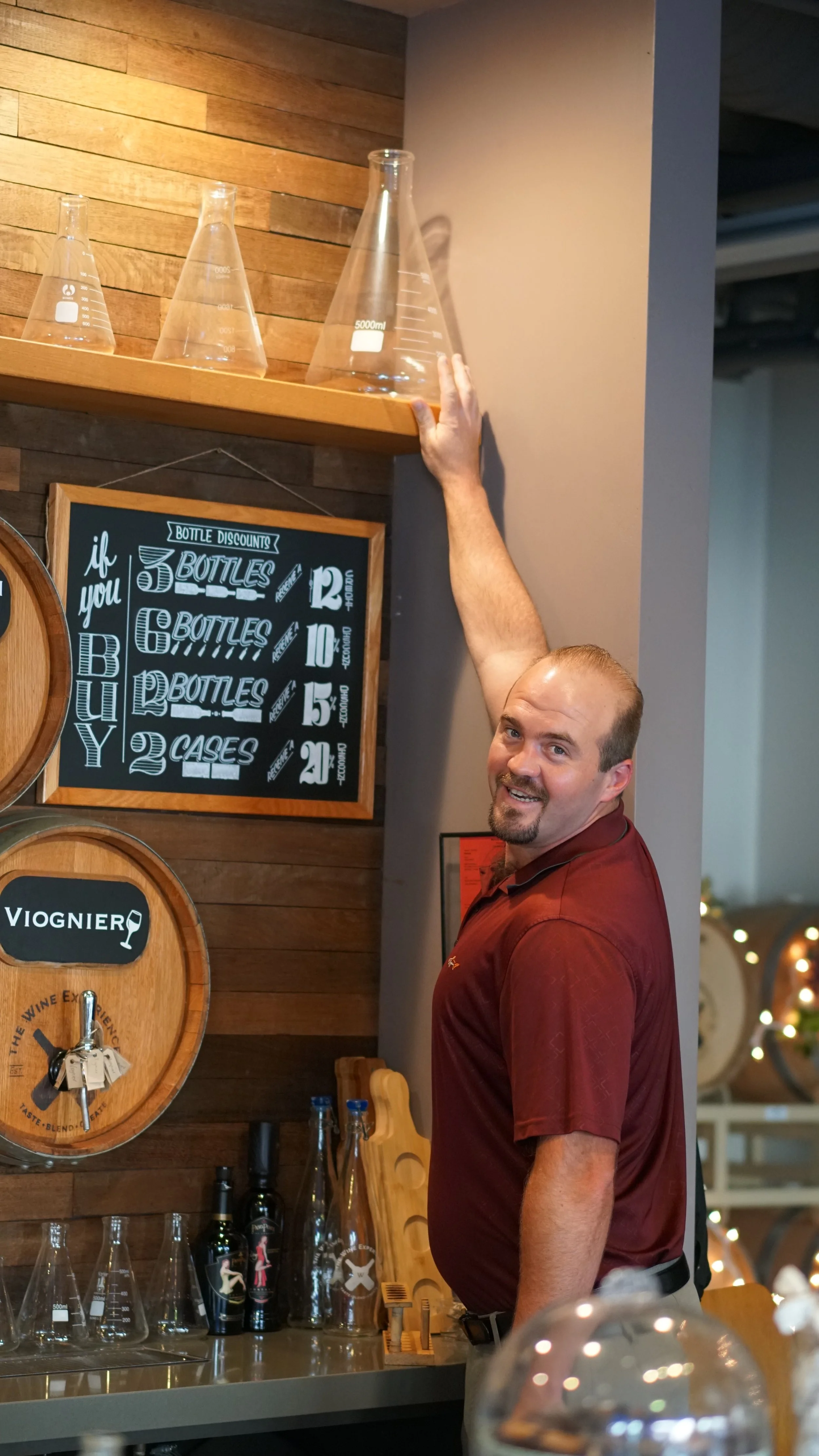 A man in a maroon polo shirt reaching up to touch a large glass beaker on a wooden shelf in a tasting room, with another smaller beakers and bottles on the wall behind him.