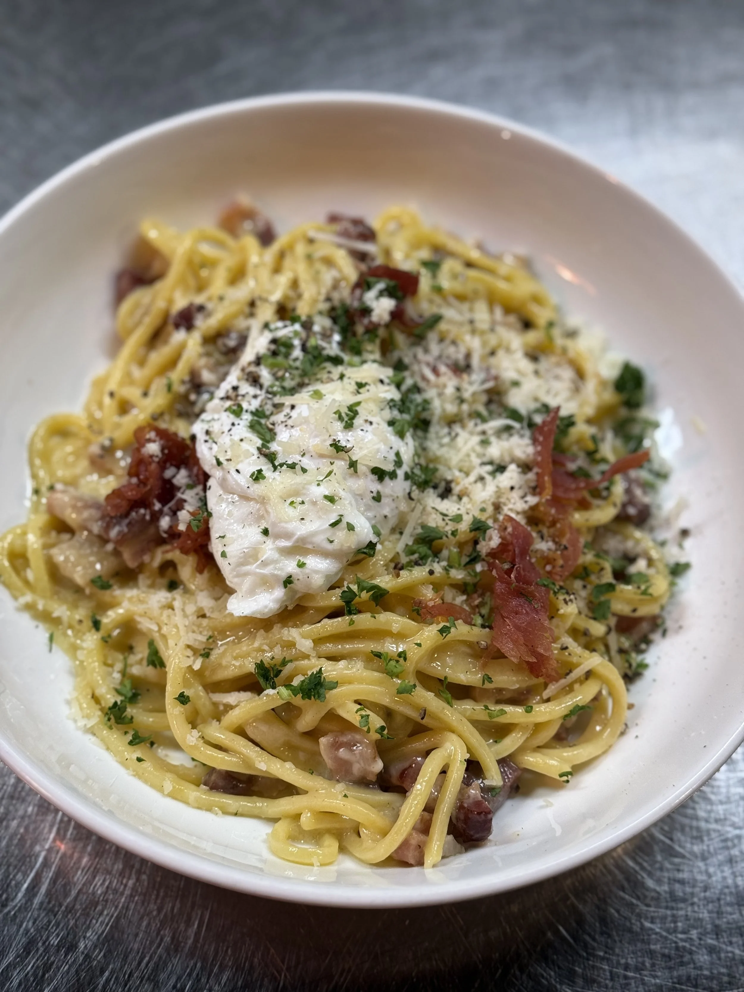 Bowl of spaghetti pasta with poached eggs, bacon, chopped parsley, and grated cheese.