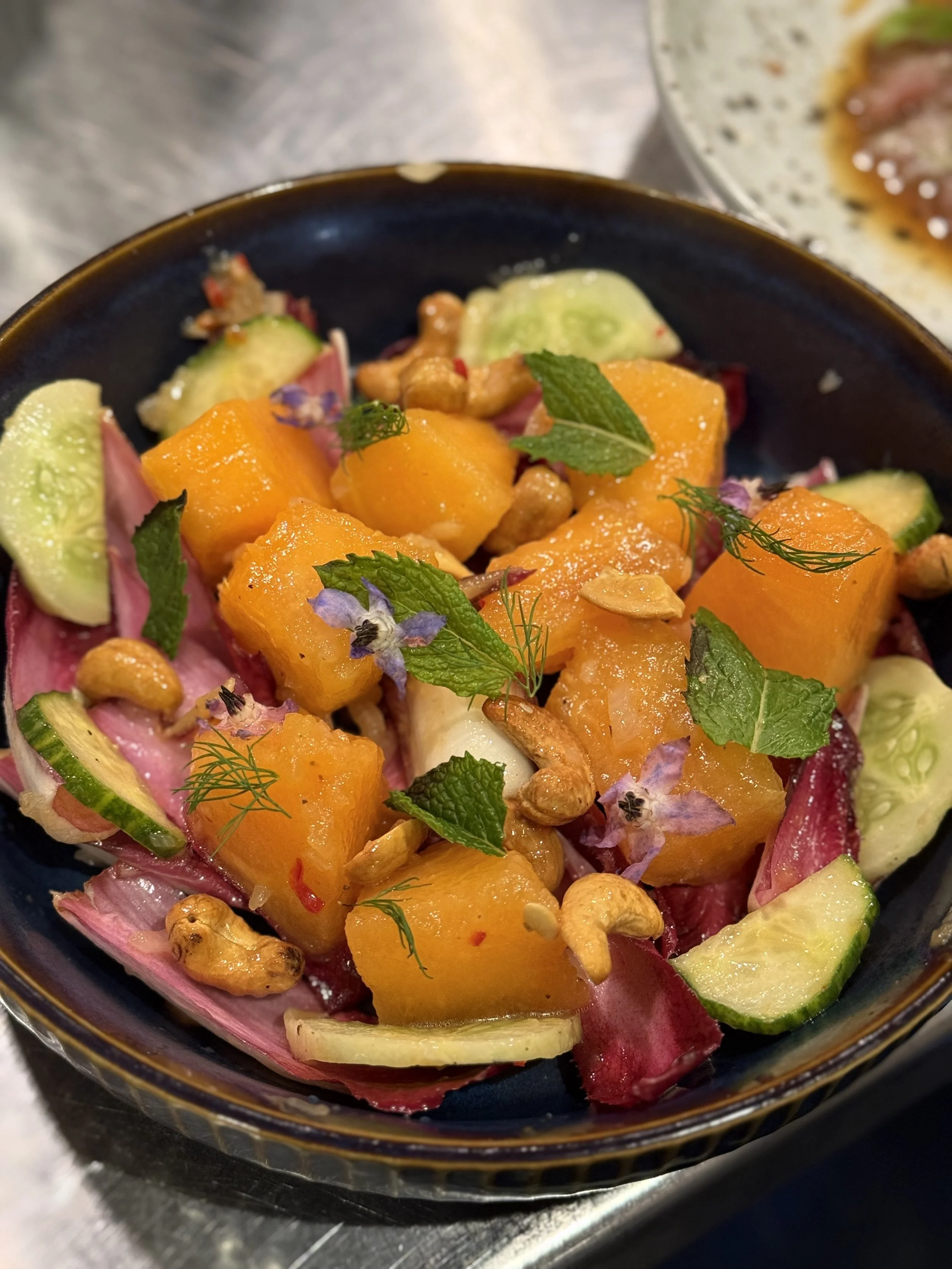 Salad with cubed orange squash, cucumber slices, red onion, chopped herbs, nuts, and edible flowers in a black bowl.