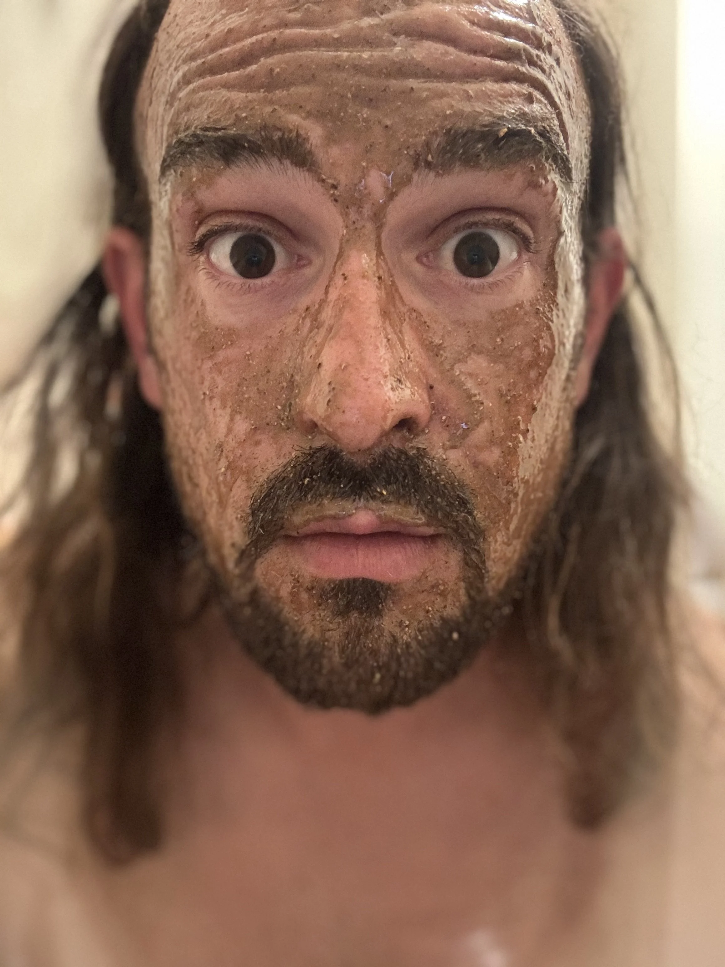 Close-up of a man with long hair, a beard, and wide eyes, covered in a brown, gritty substance on his face, looking surprised or startled. The mud mask on his face looks textured and natural filled with a combination of ingredients.