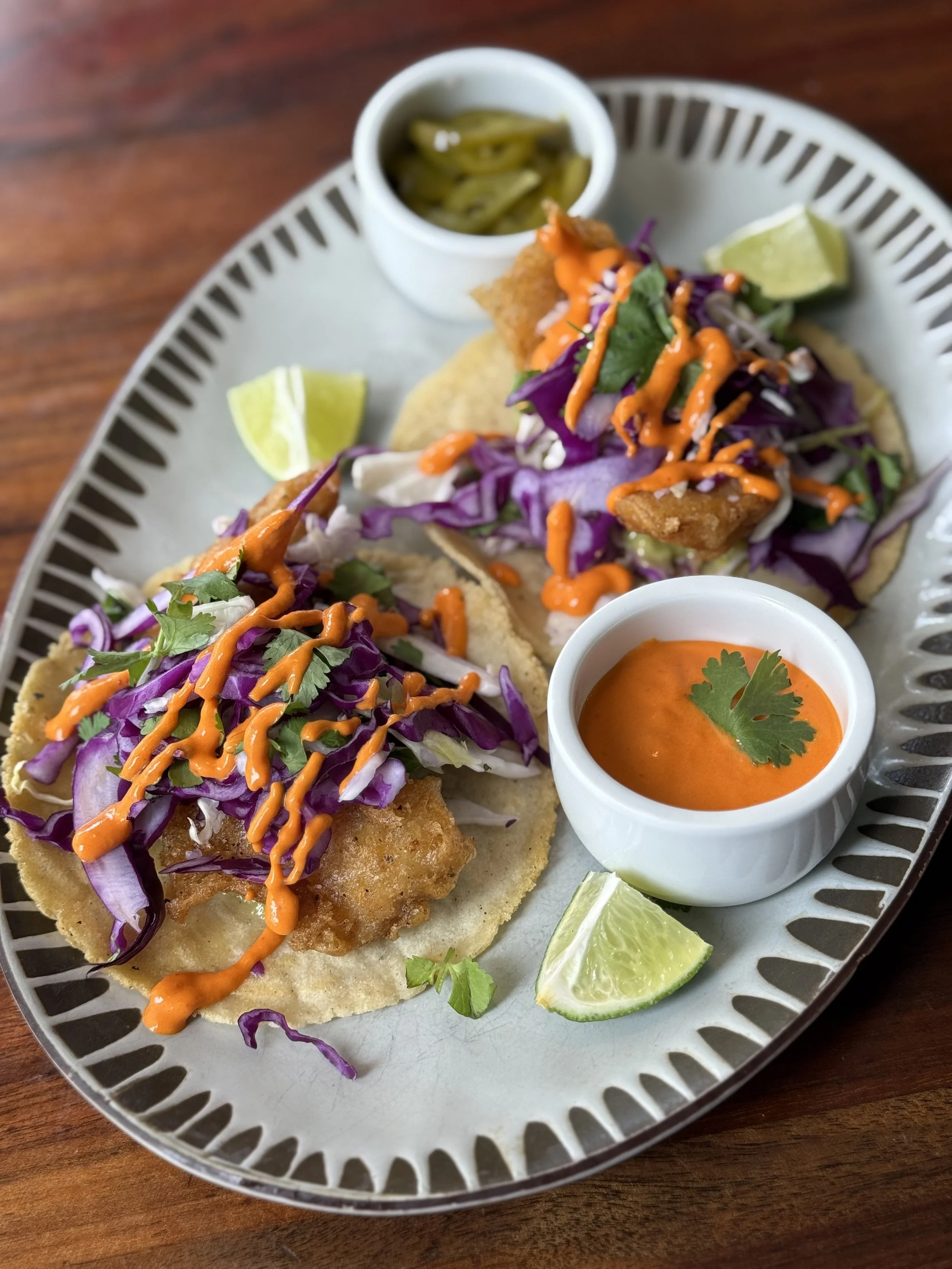 Two fish tacos topped with purple cabbage slaw and orange sauce, served with lime wedges, a small bowl of red sauce garnished with cilantro, and another small bowl of pickled jalapeños on a decorative white plate.