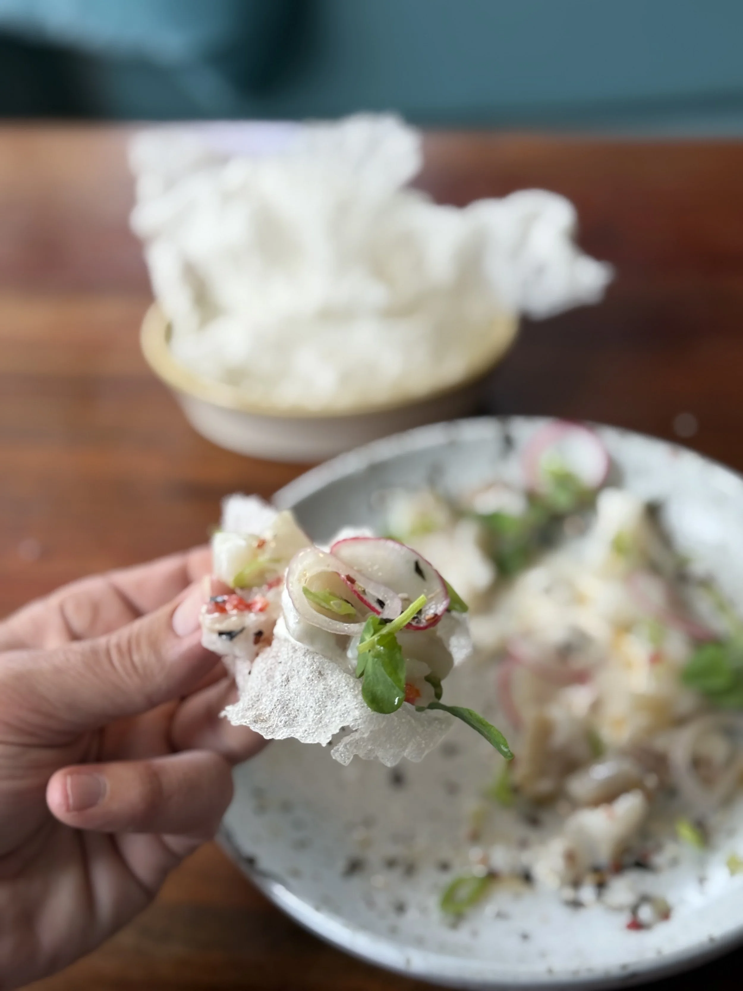 Hand holding a rice paper roll filled with sliced vegetables, garnished with green herbs, with a bowl of more rice paper rolls and a bowl of rice crackers in the background.